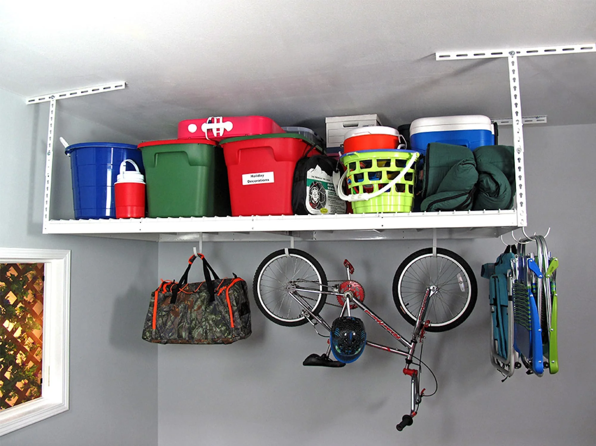 Garage shelf holding various items including large storage bins, a cooler, a laundry basket, a black bag, a camouflage duffel bag, a bicycle, a detachable helmet, and a rolling seat, with a wall, a window with a lattice pattern, and part of a wall-mounted rack visible.