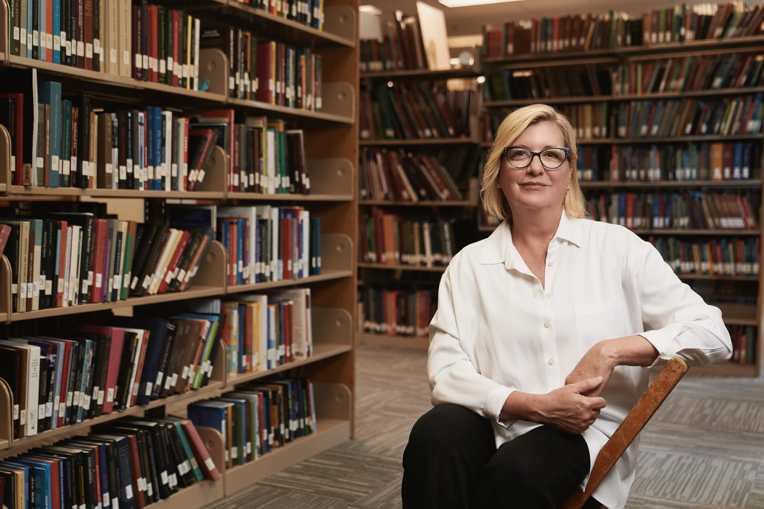 A woman with blonde hair, glasses, and a white shirt sitting in a library next to bookshelves filled with colorful books, holding a book.