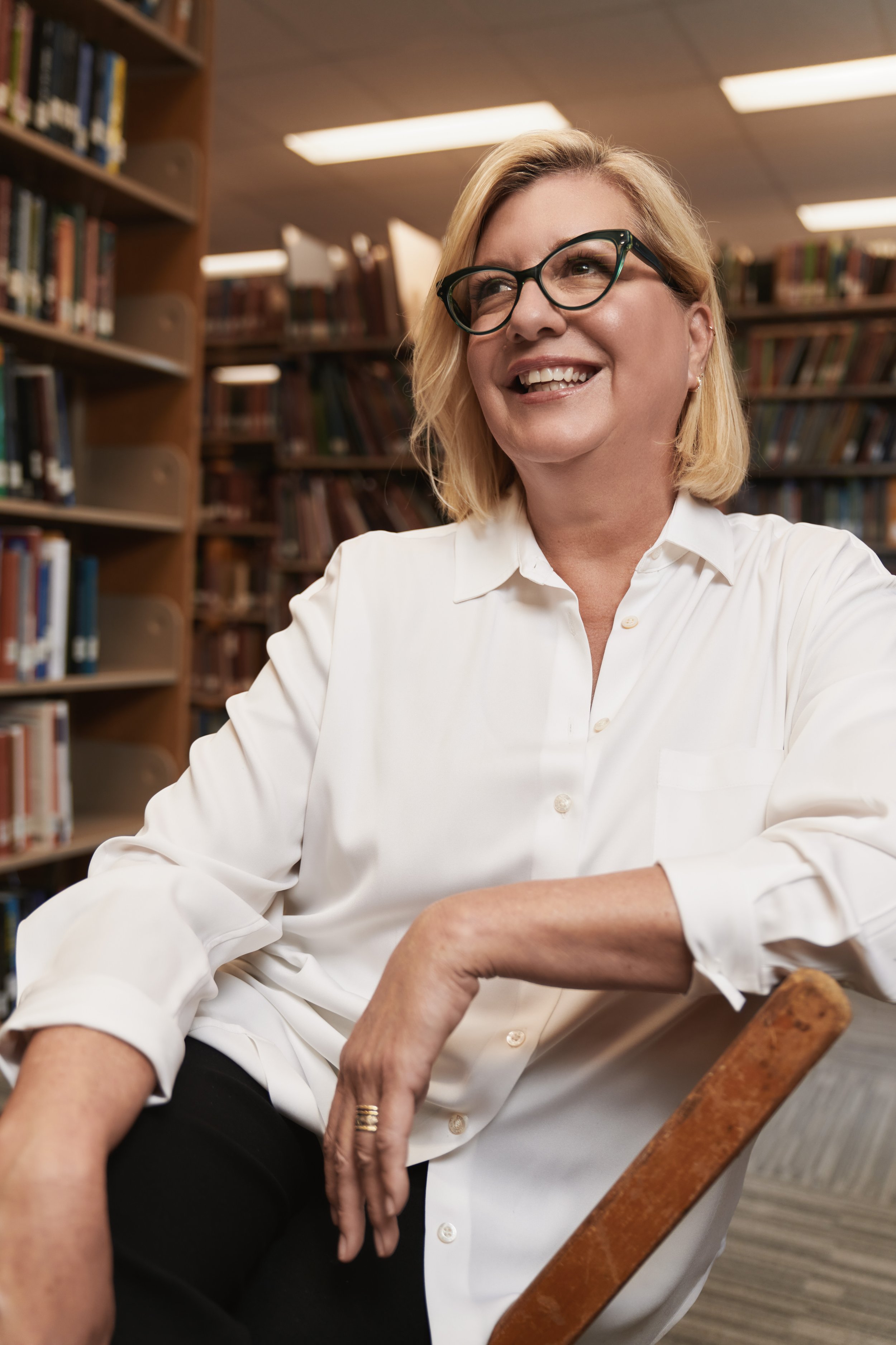 A woman with blonde hair, glasses, and a white shirt sitting in a library with bookshelves behind her, smiling and looking to the side.