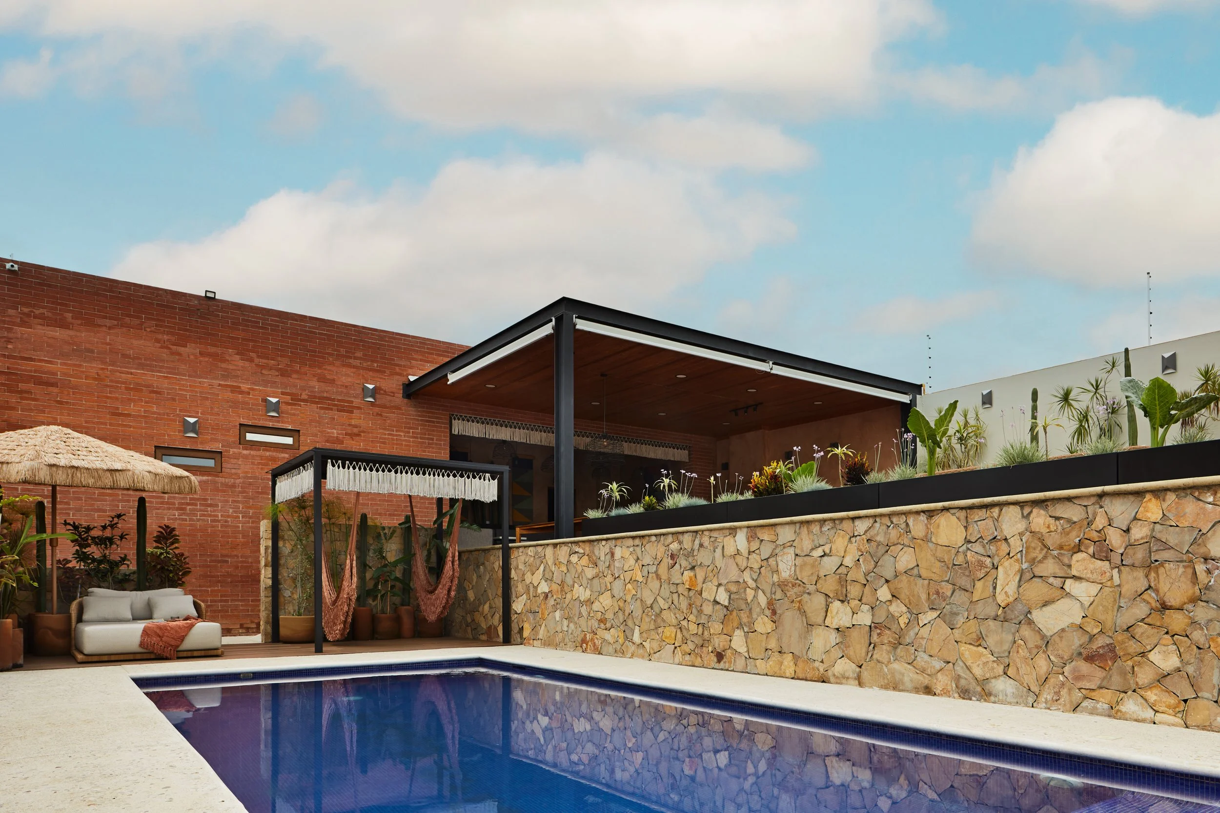 Vista de una moderna área de piscina exterior con mobiliario cómodo, plantas y un techo cubierto, contra un muro de ladrillo y un cielo con nubes.