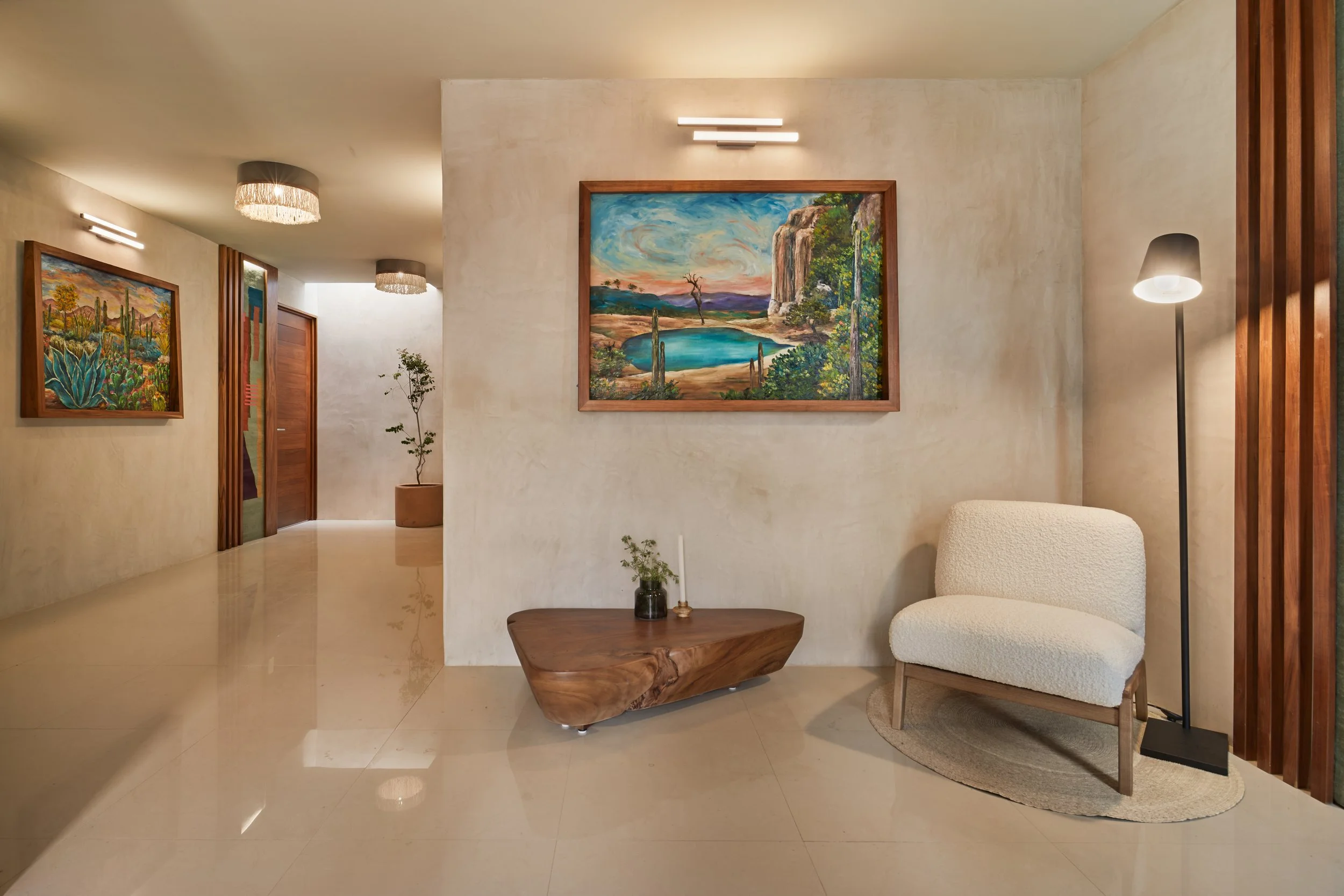 Sala de estar moderna con paredes beige, cuadros enmarcados con paisajes y vegetación. Muebles incluyendo una silla blanca, mesa de madera y una lámpara de piso negra. Iluminación moderna en el techo y lámparas ambientales.