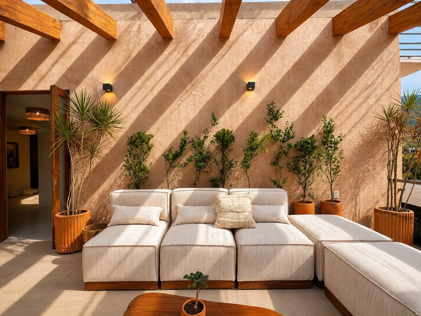 Sala de estar con sillones blancos, cojines, plantas en macetas grandes y pequeñas, pared de textura beige con sombra y ventanales