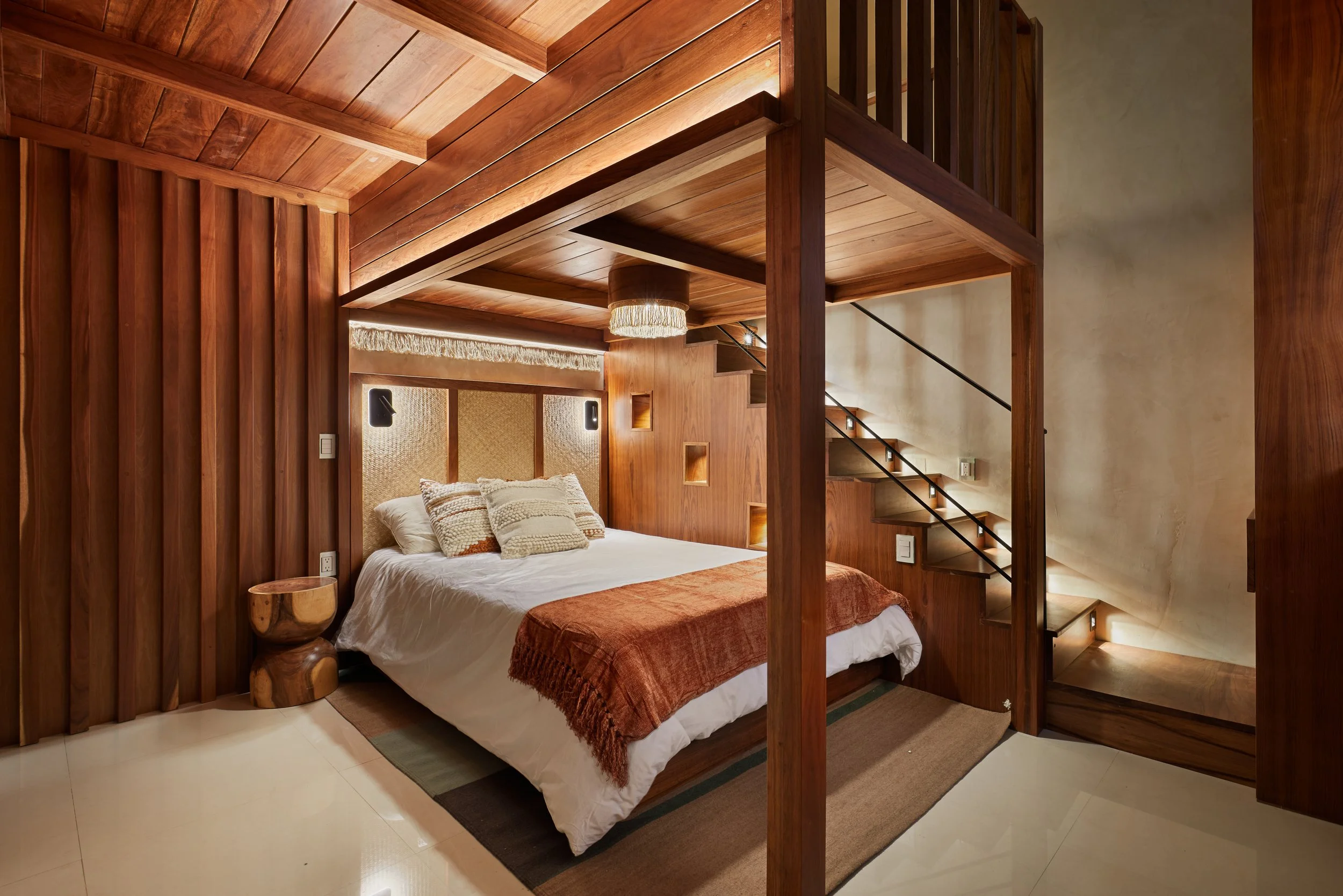 Habitación con estructura de madera, cama con ropa blanca y decoraciones en tonos cálidos, lámpara en el techo y escalera de madera