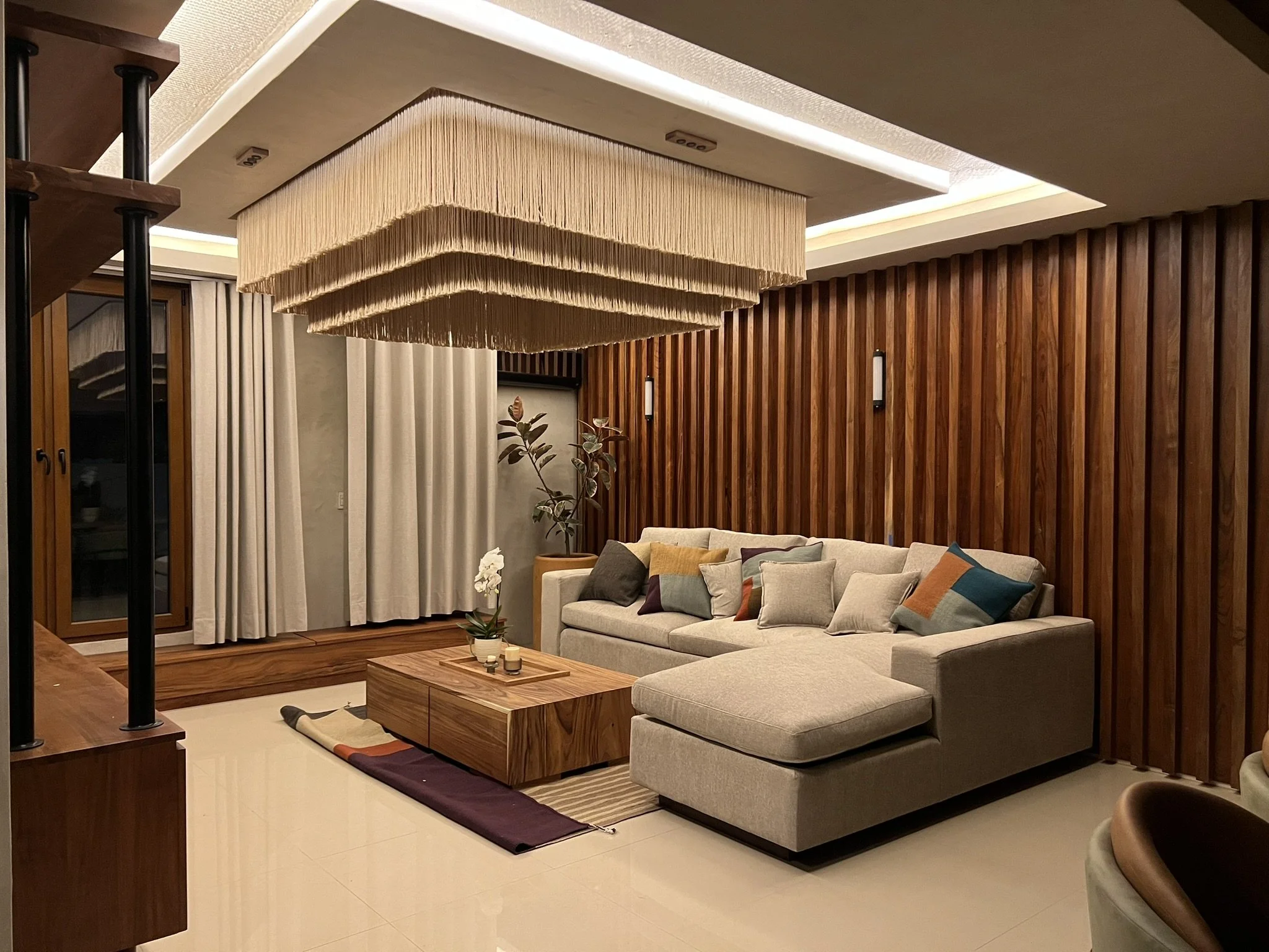 Sala moderna con paredes de madera, lámparas y cortinas blancas, con sillones y una mesa de centro decorada con velas y plantas.