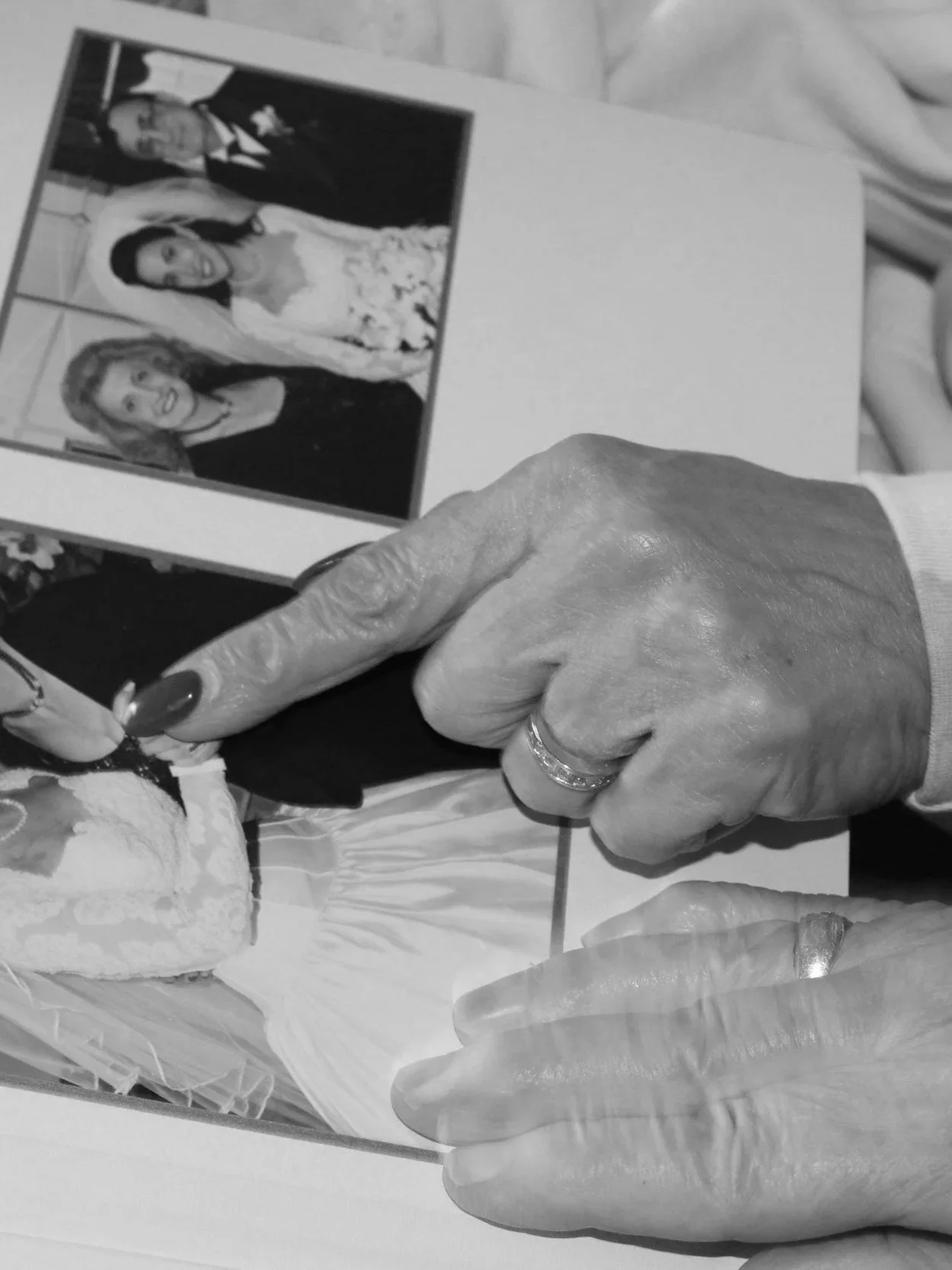 Close-up of an elderly woman's hand pointing at a wedding photo of a young bride in a wedding dress with two other people, on a table.