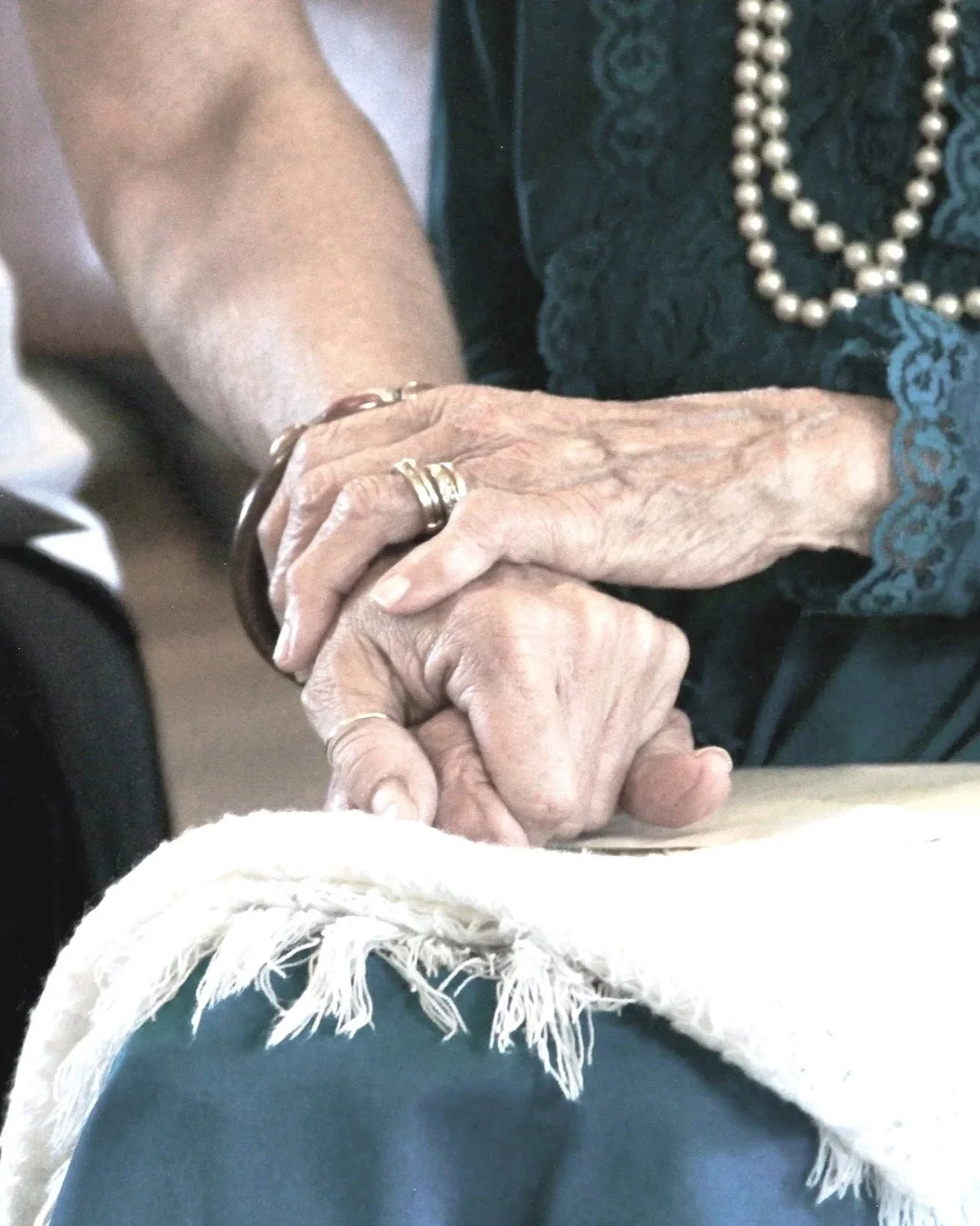 Close-up of elderly hands intertwined, resting on a cloth-covered surface. The person is wearing a dark shirt with blue embroidery, a pearl necklace, and multiple rings.