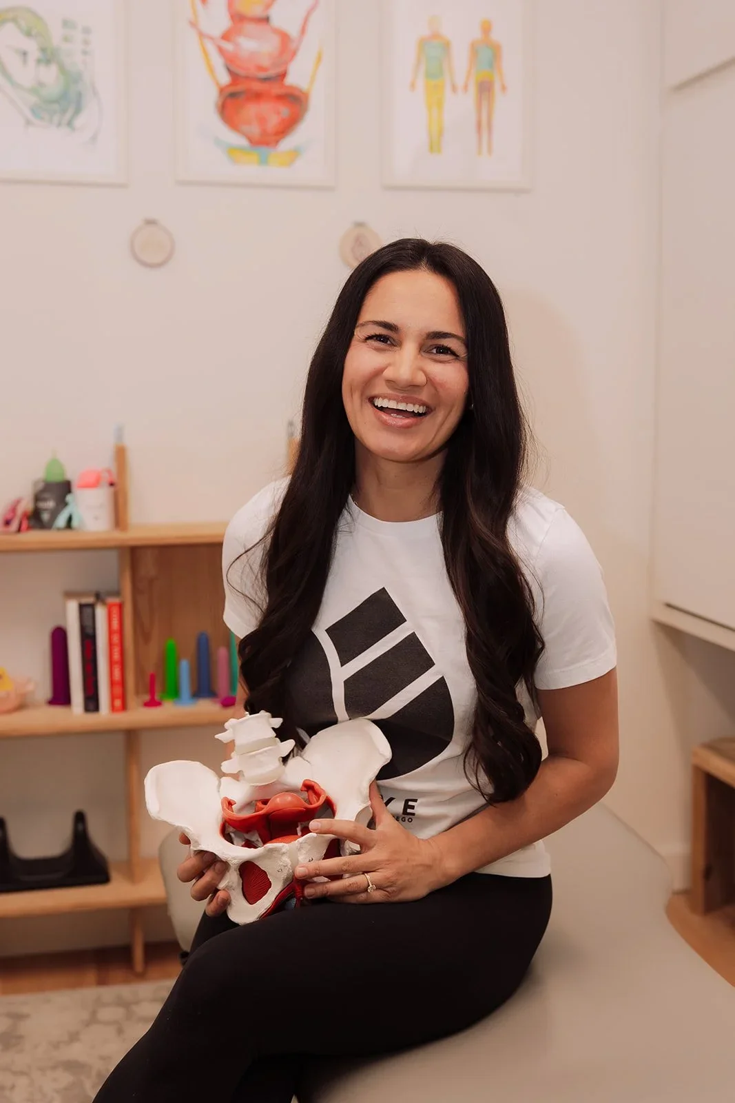 A smiling woman with long dark hair holding a pelvic anatomy model, sitting in a room with medical posters and toys in the background.