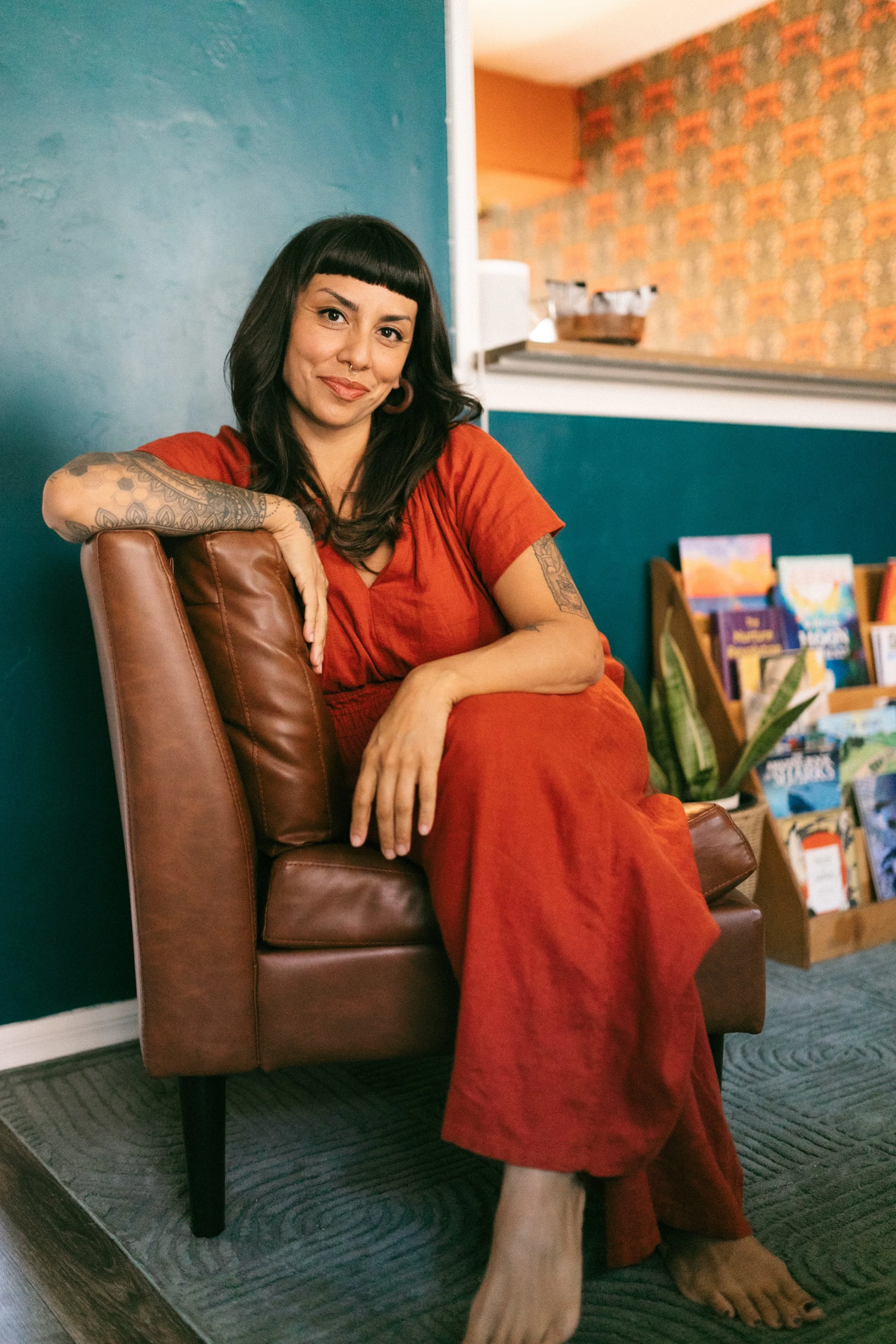 A woman with dark hair and tattoos, wearing a red jumpsuit, sitting on a leather chair in a colorful room with bookshelves.