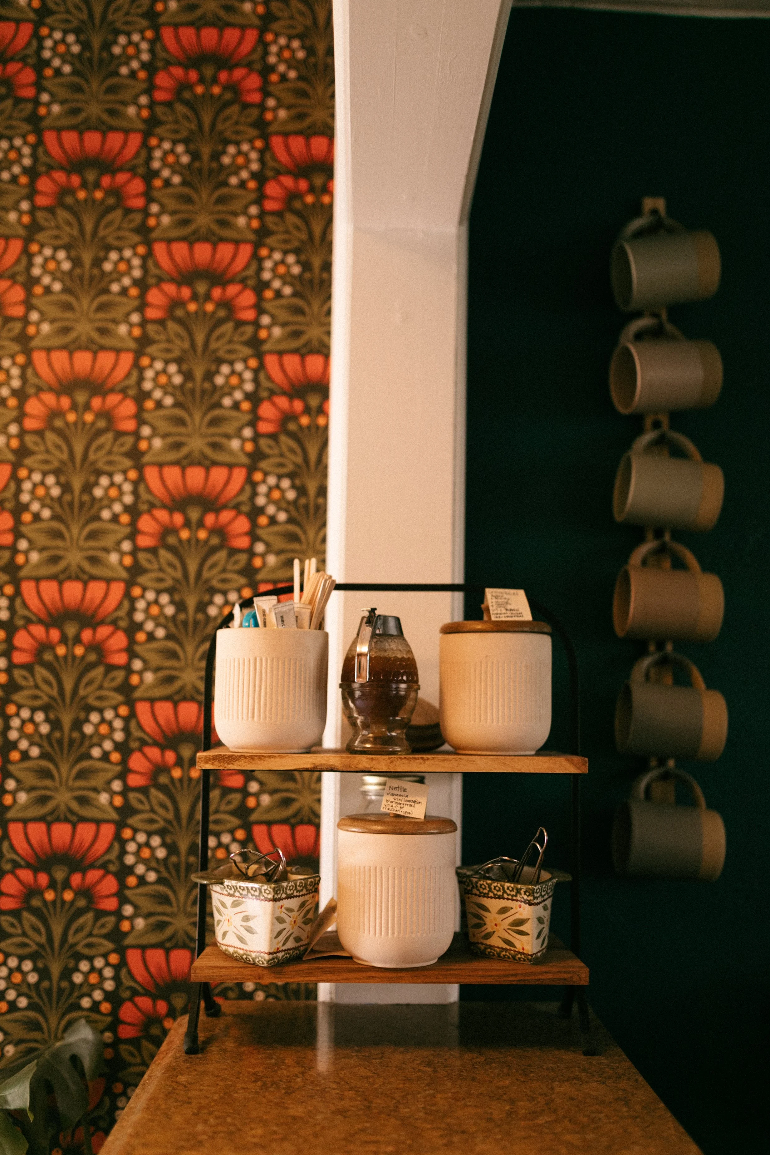 Decorative display with white ceramic containers holding utensils, on a wooden stand, against a patterned wallpaper with orange and green floral designs.