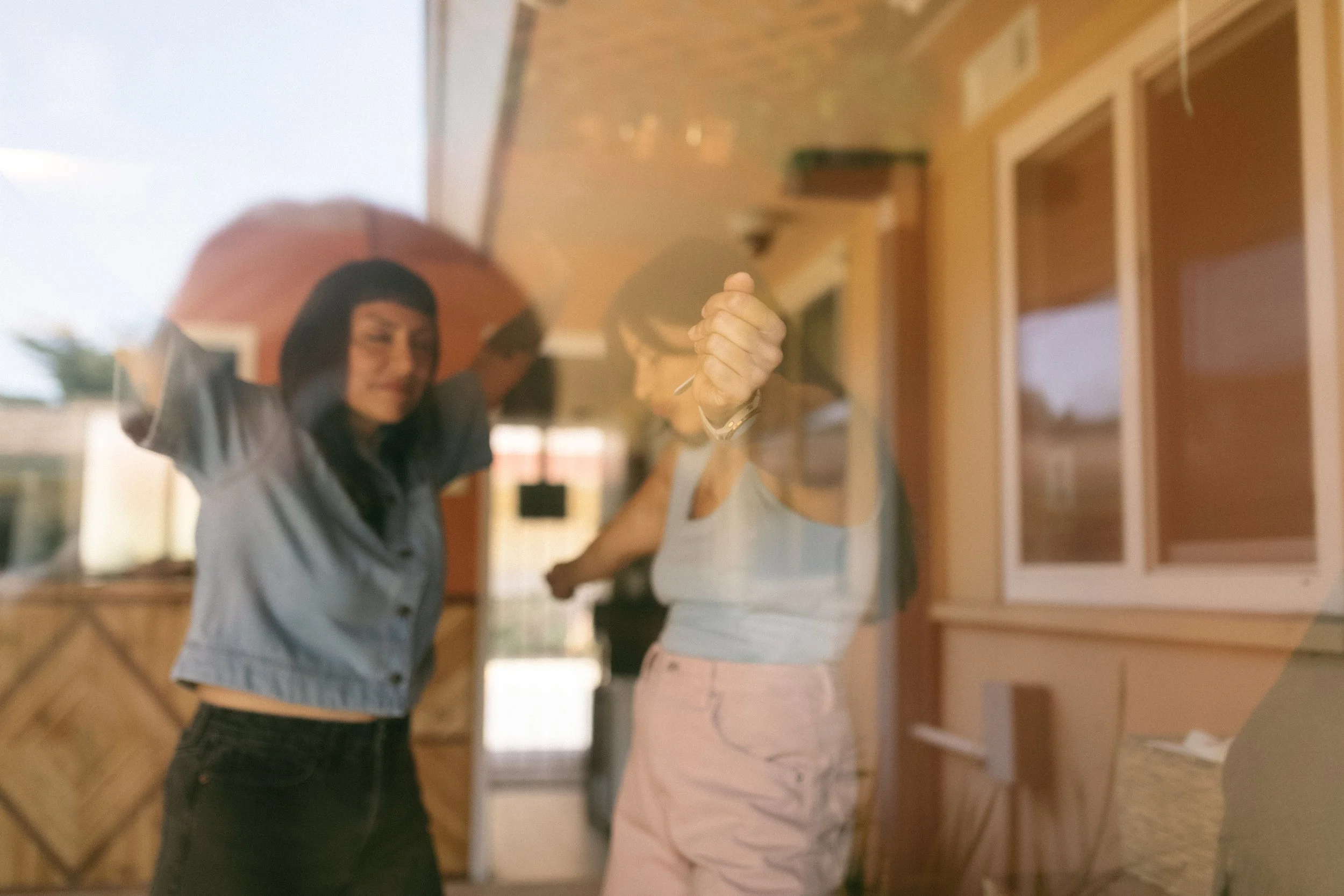 Through a window, a woman with black hair wearing a blue shirt and black pants dancing with another woman with black hair, gray shirt, and pink pants.