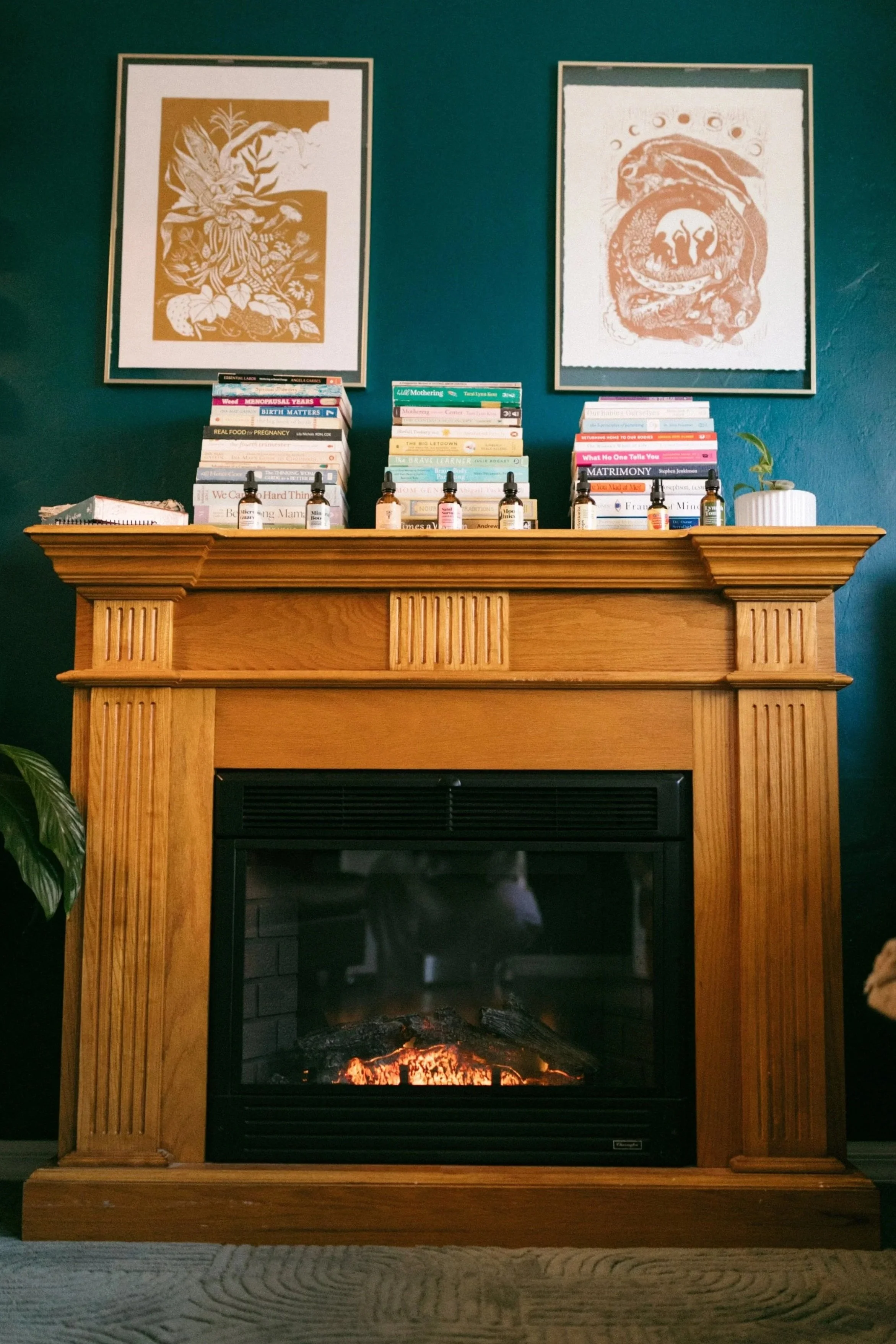Wood fireplace with a burning fire, topped with pictures of books and bottles of essential oils on the mantel, against a dark teal wall decorated with two framed art prints.