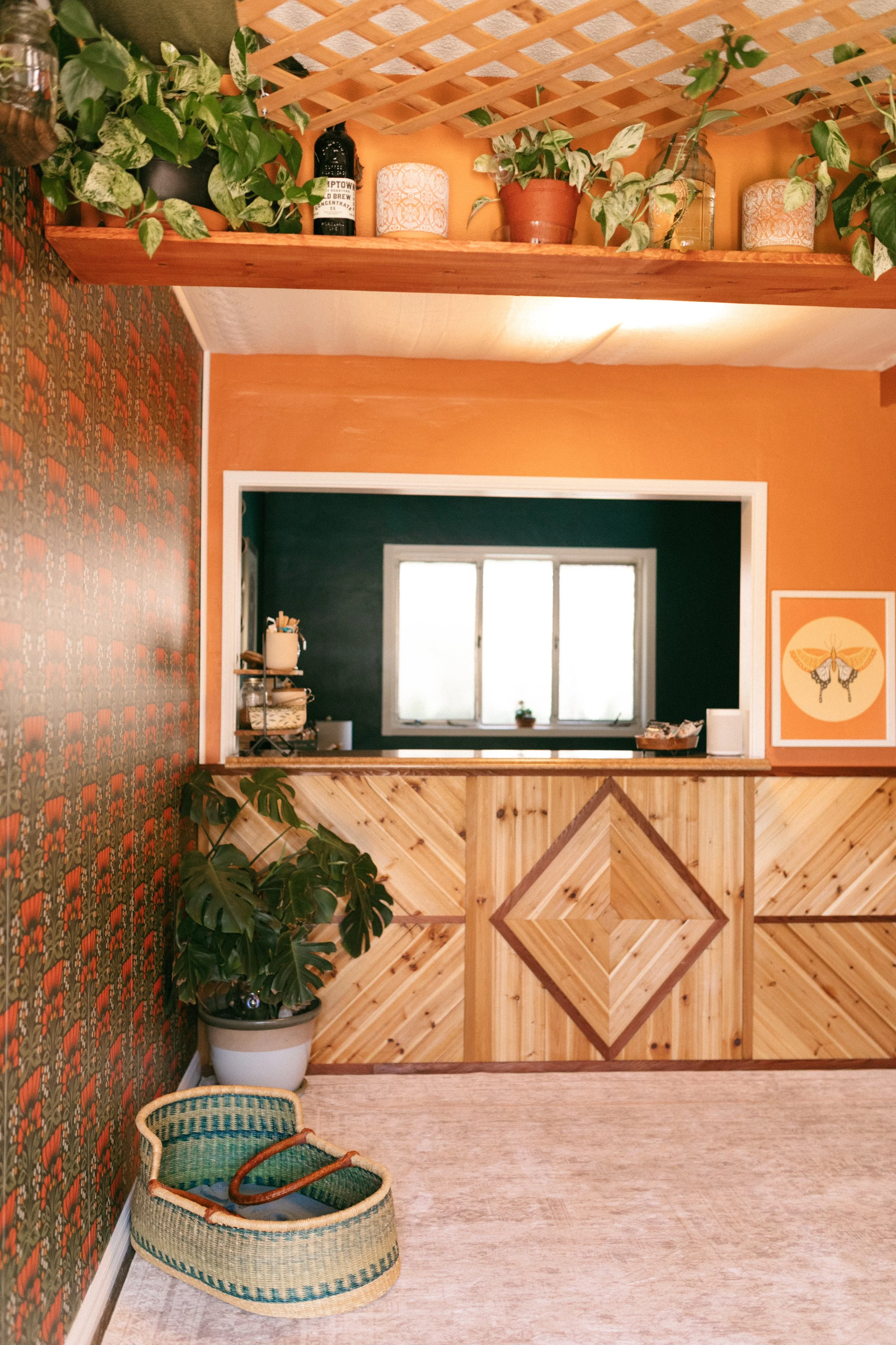 Interior view of a cozy room with a wooden counter, a window, potted plants on a shelf, patterned wallpaper, and a woven basket on the floor.