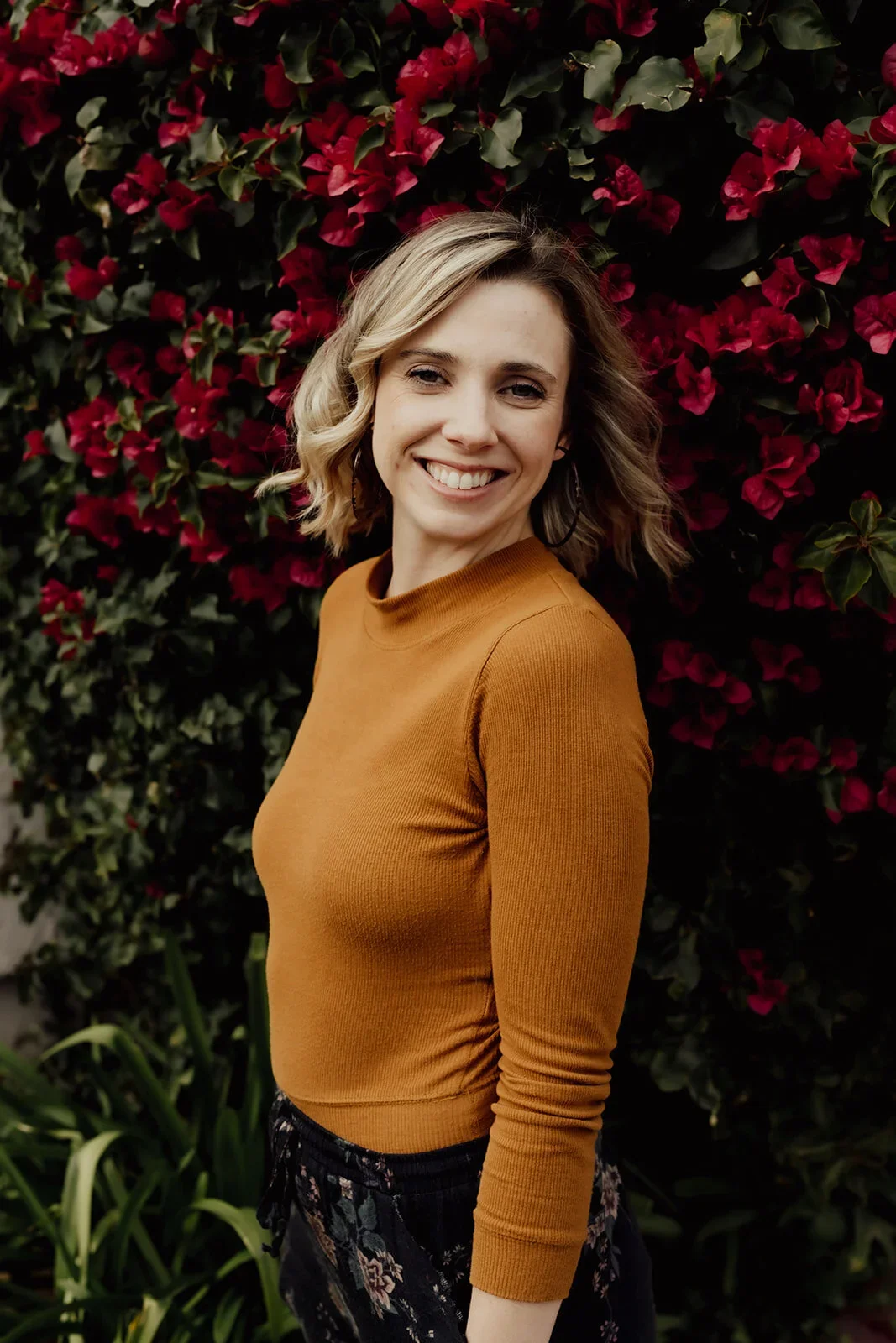 A woman with shoulder-length blonde hair smiling outdoors in front of a large bush of pink flowers, wearing a mustard-colored long sleeve shirt.