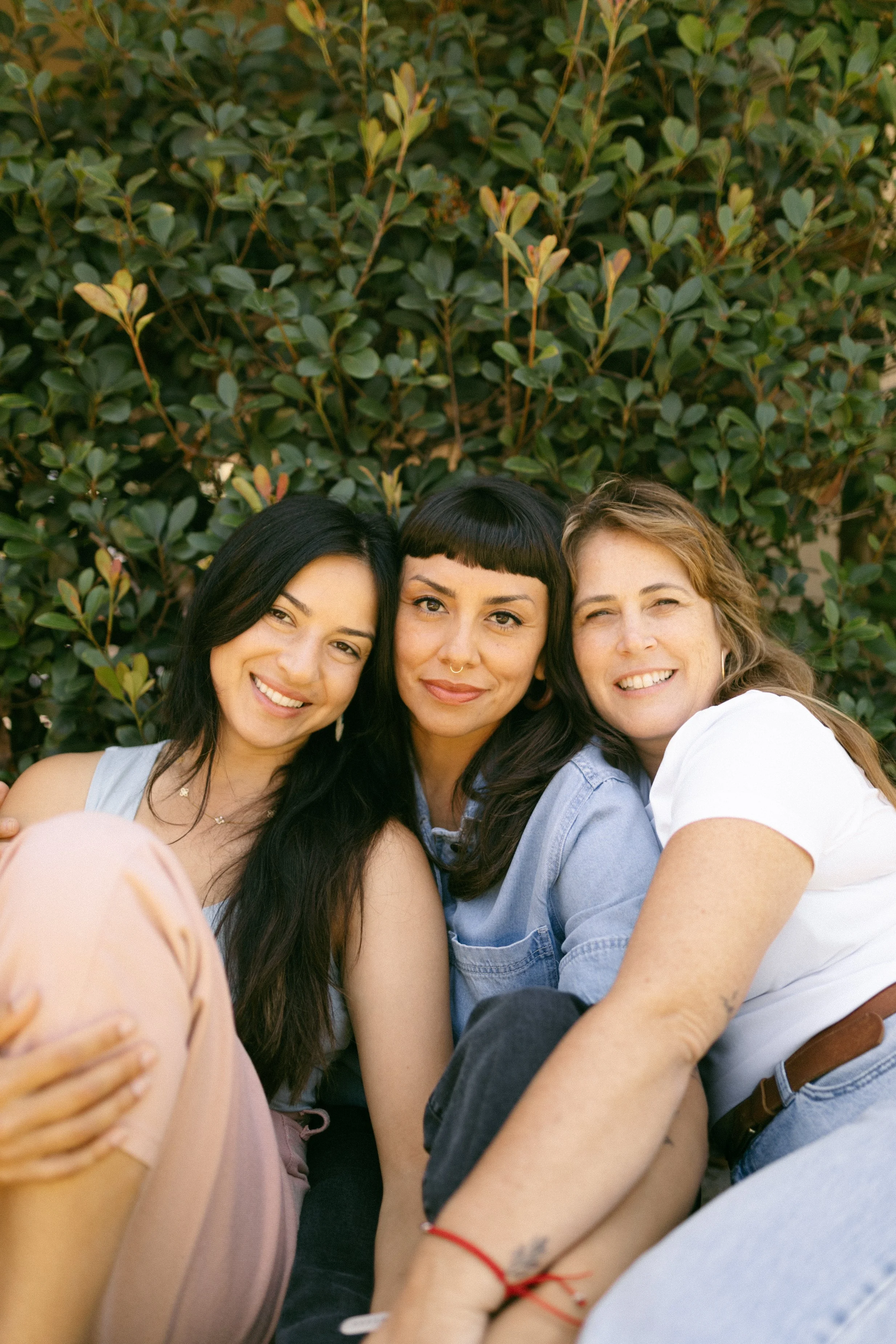 Group of three women sitting close together outdoors, with green leafy shrubbery in the background, smiling.