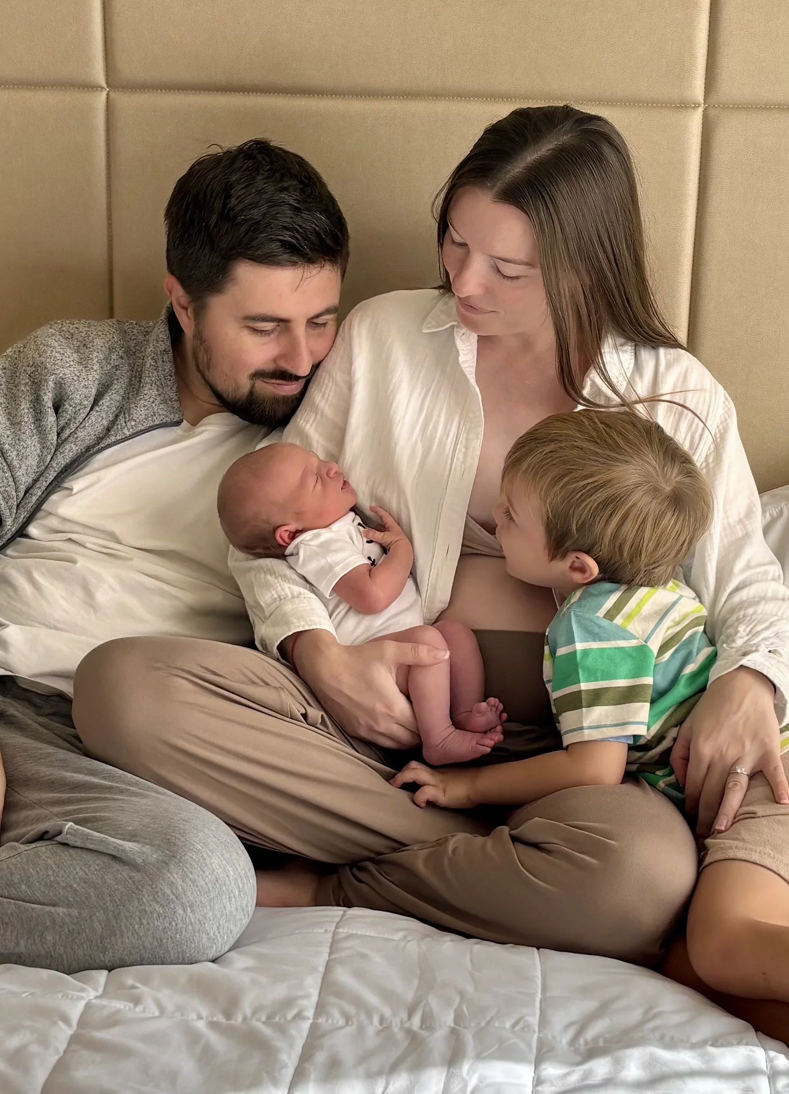 A family of four sitting on a bed, with a newborn baby and an older child, all looking lovingly at the baby.