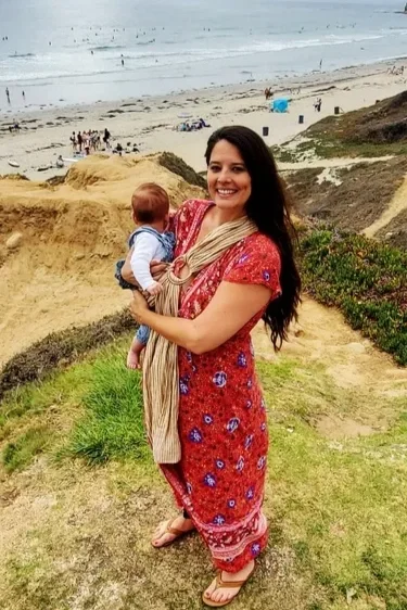 Woman in a red patterned dress holding a baby near a beach with cliffs and ocean in the background.