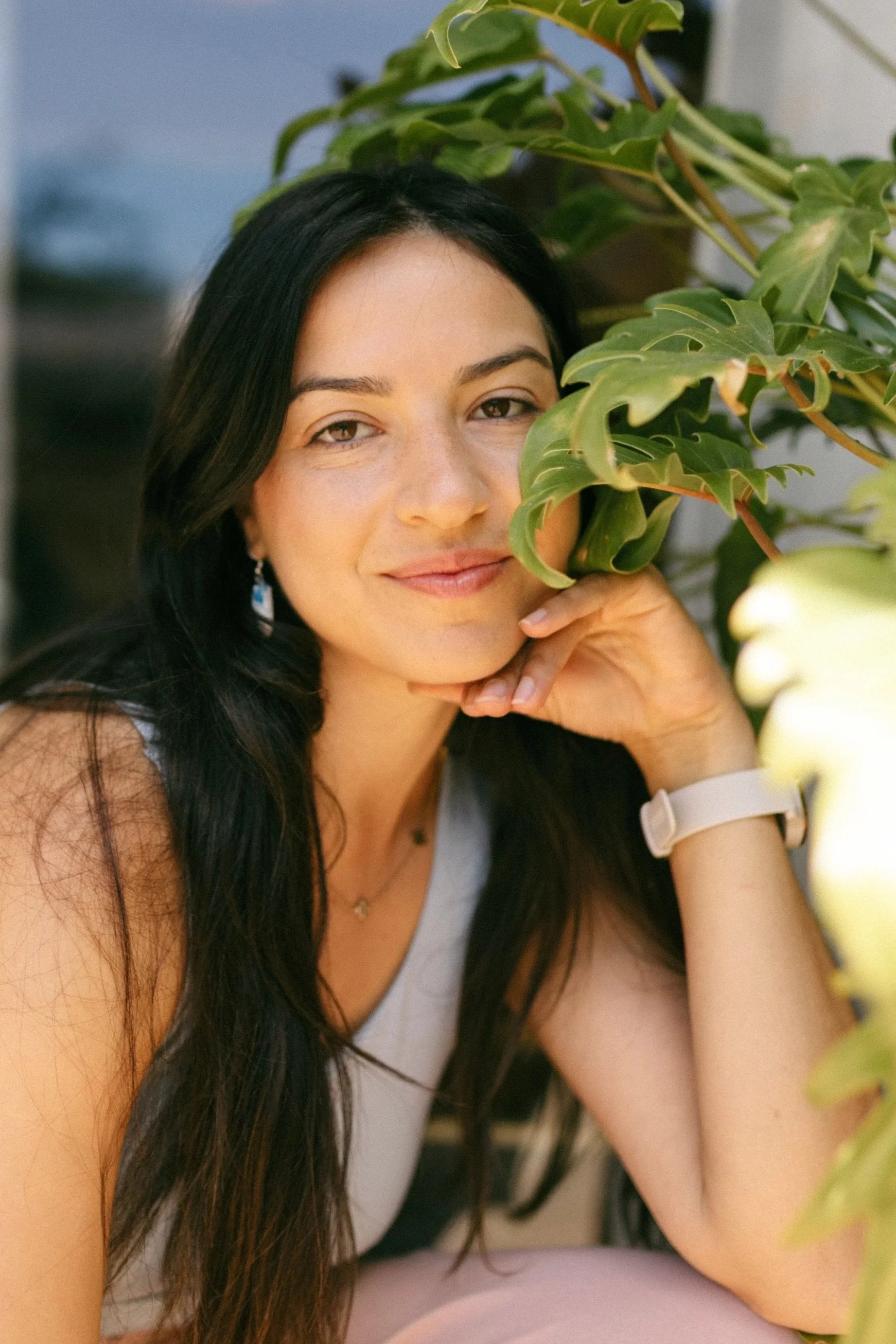 A woman with long dark hair, wearing a white wristwatch and a light-colored tank top, sitting in front of a green leafy plant, smiling gently and resting her chin on her hand.