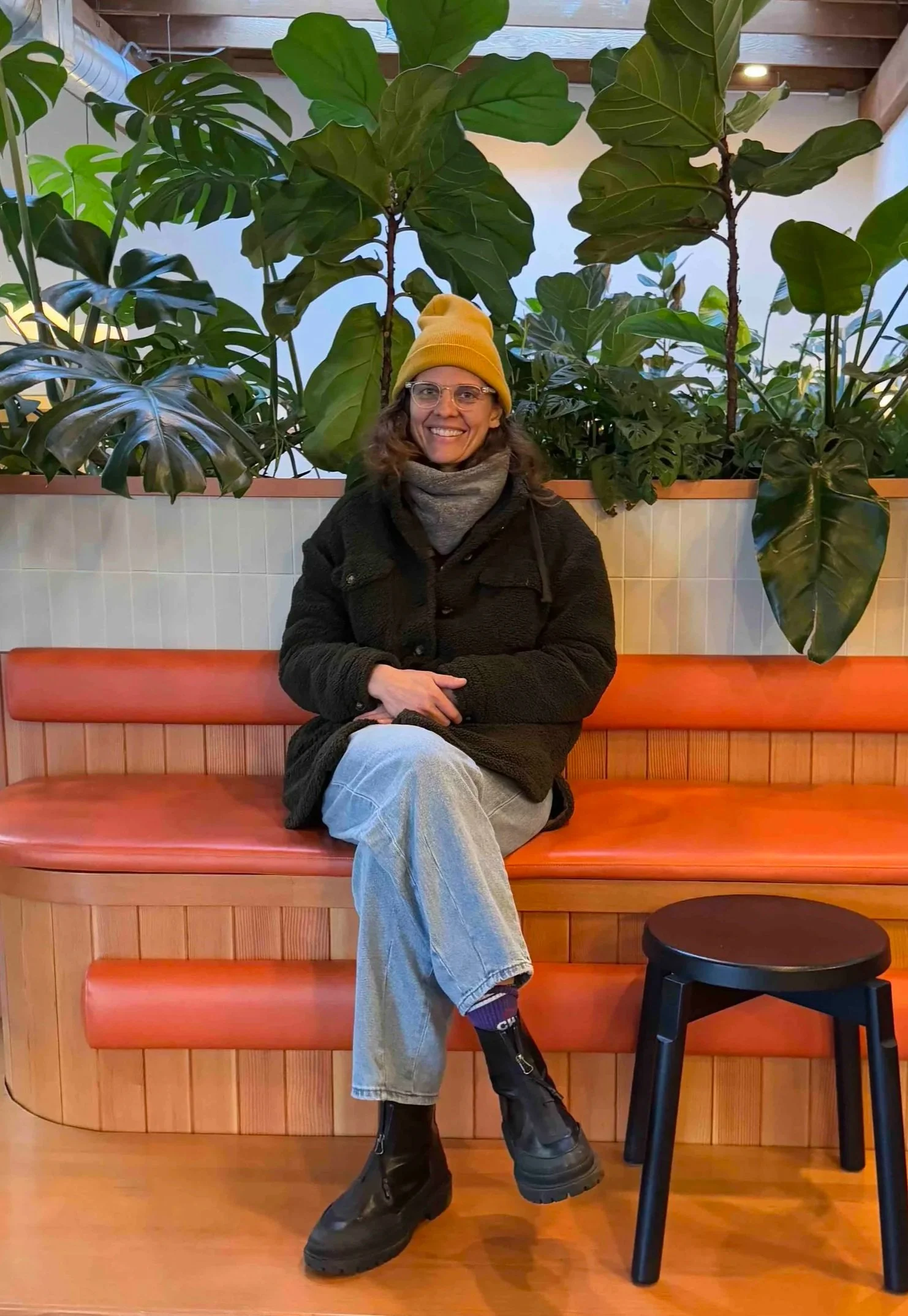 A woman with glasses, a yellow beanie, black coat, gray scarf, light gray pants, and black boots sitting on a wooden bench in front of green leafy plants. Nutritionist, Dietitian, Portland Oregon