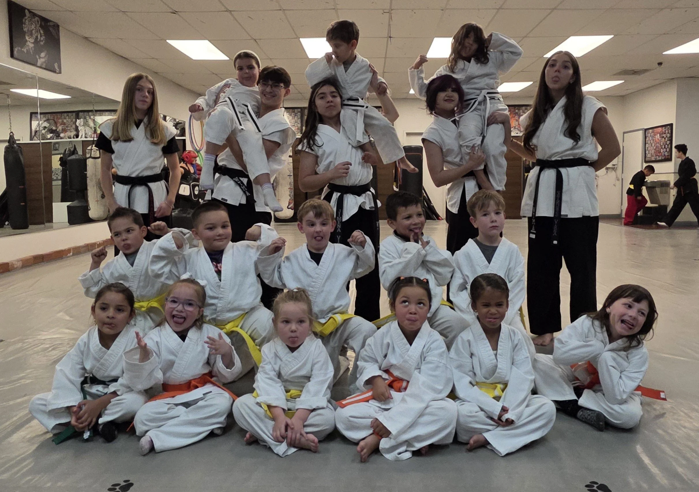 Group of children and teenagers in martial arts uniforms posing in a dojo, some standing and some sitting on the floor, with instructors in the background.