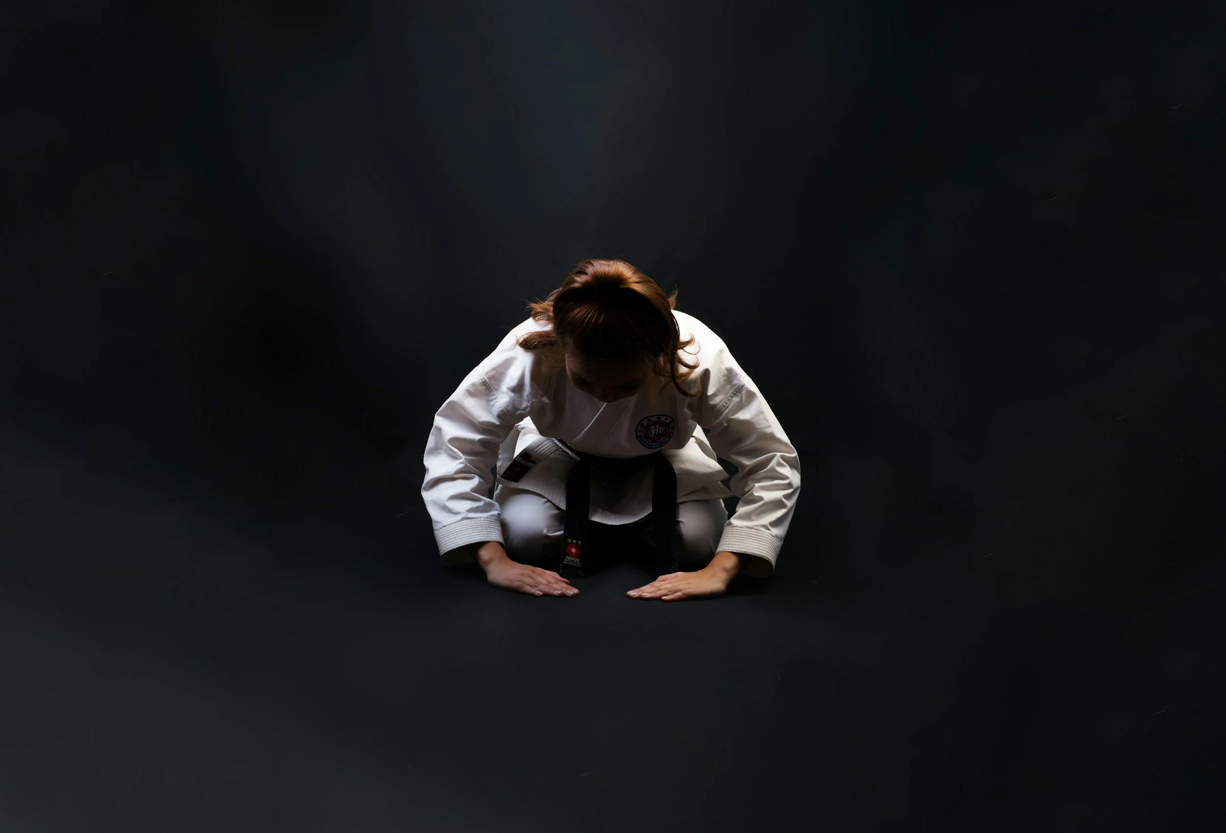 A woman in a white martial arts uniform with a black belt is kneeling on the ground, bowing her head and touching her hands to the floor in a gesture of respect, against a dark background.