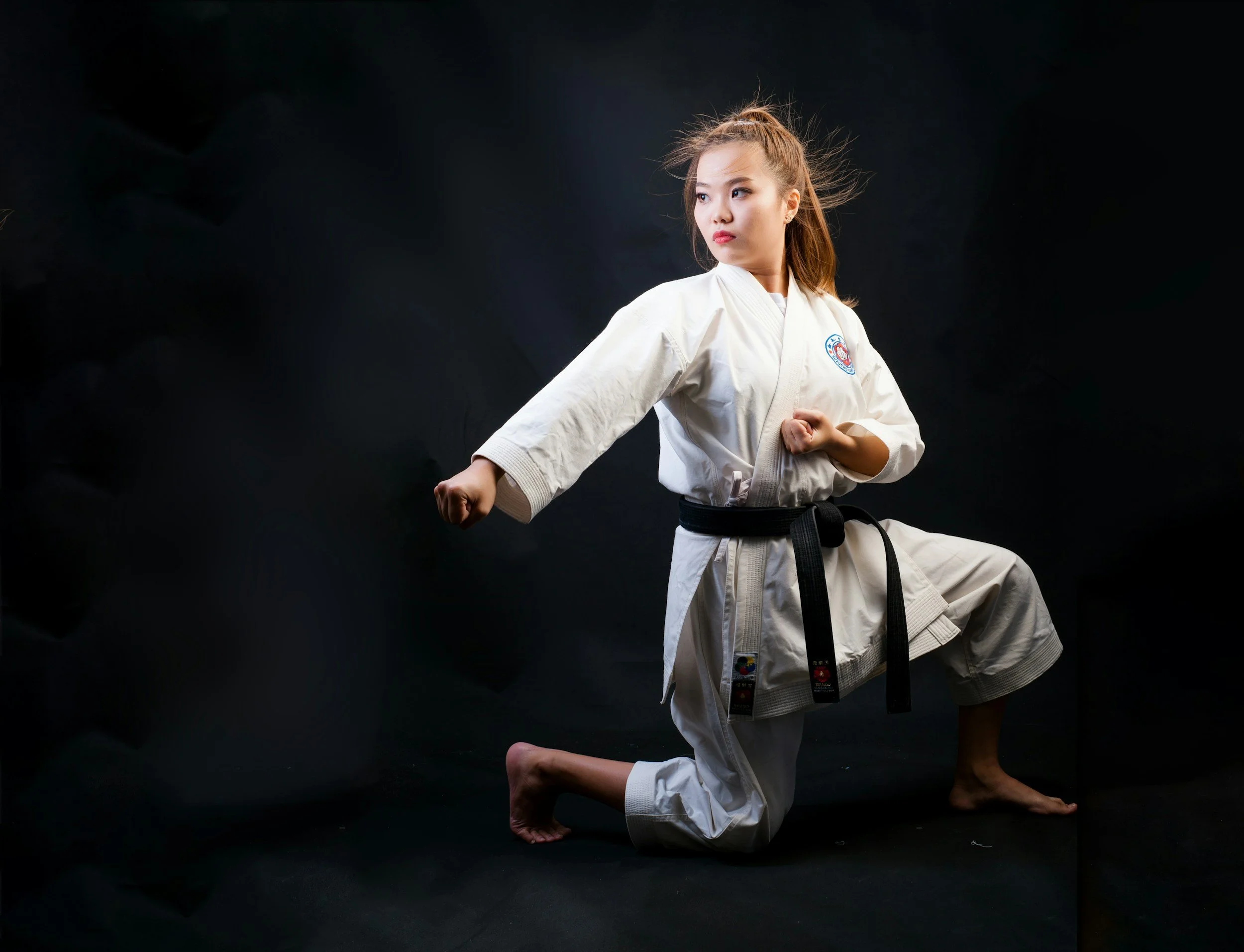 A young woman in a white martial arts gi performing a high side kick against a black background.