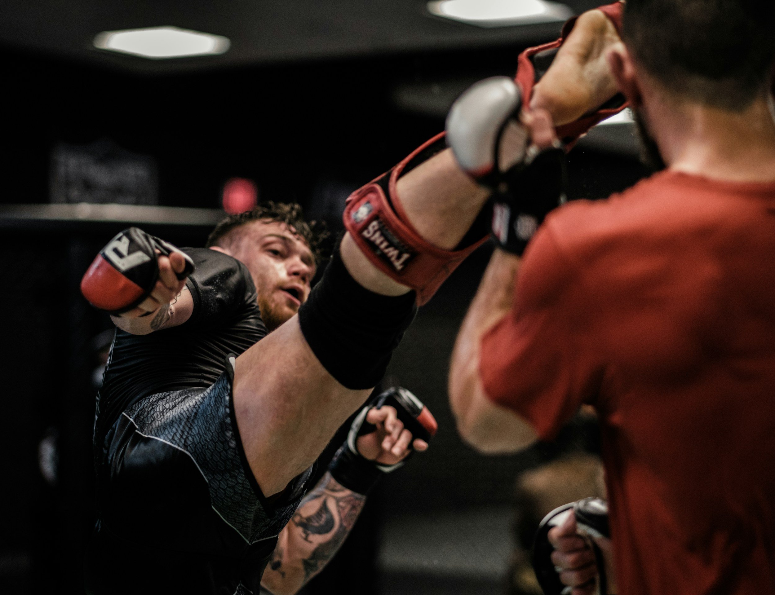 Two male fighters engaged in a mixed martial arts fight, wearing gloves, one in black and the other in red, inside a cage or ring, with one fighter delivering a punch to the other.