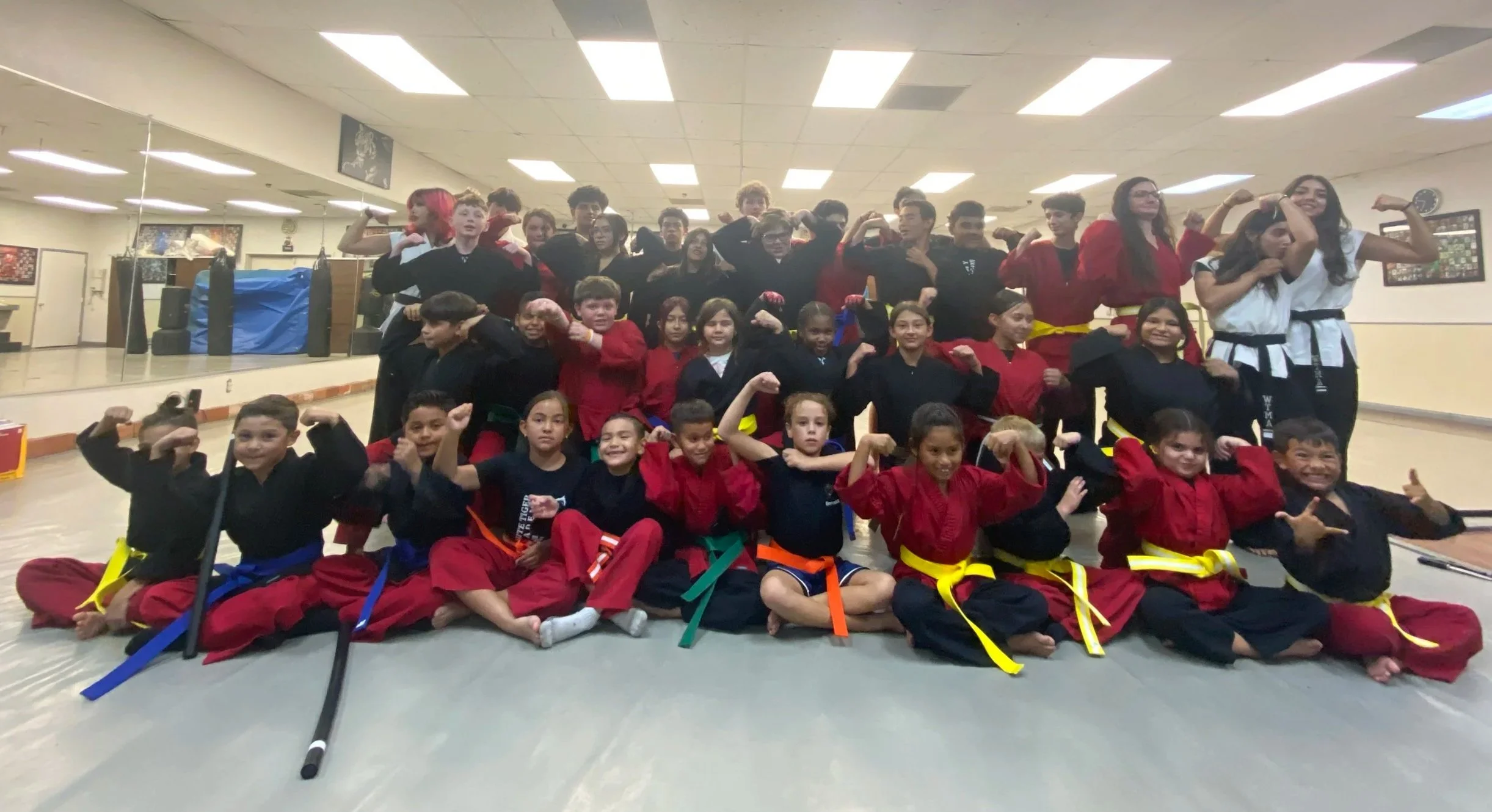Group of children and teenagers in martial arts uniforms and casual clothes, posing with flexed biceps in a martial arts studio.