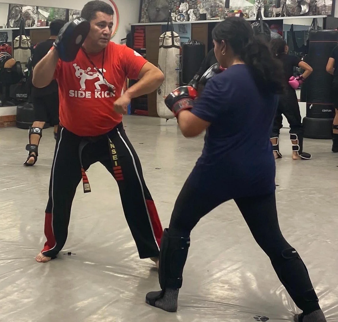 Two people training in a boxing or martial arts gym. The man on the left is wearing black pants, a red shirt, and boxing gloves, holding focus mitts. The woman on the right is wearing black leggings, a blue shirt, and boxing gloves, positioned for a punch. Several other people and boxing equipment are visible in the background.