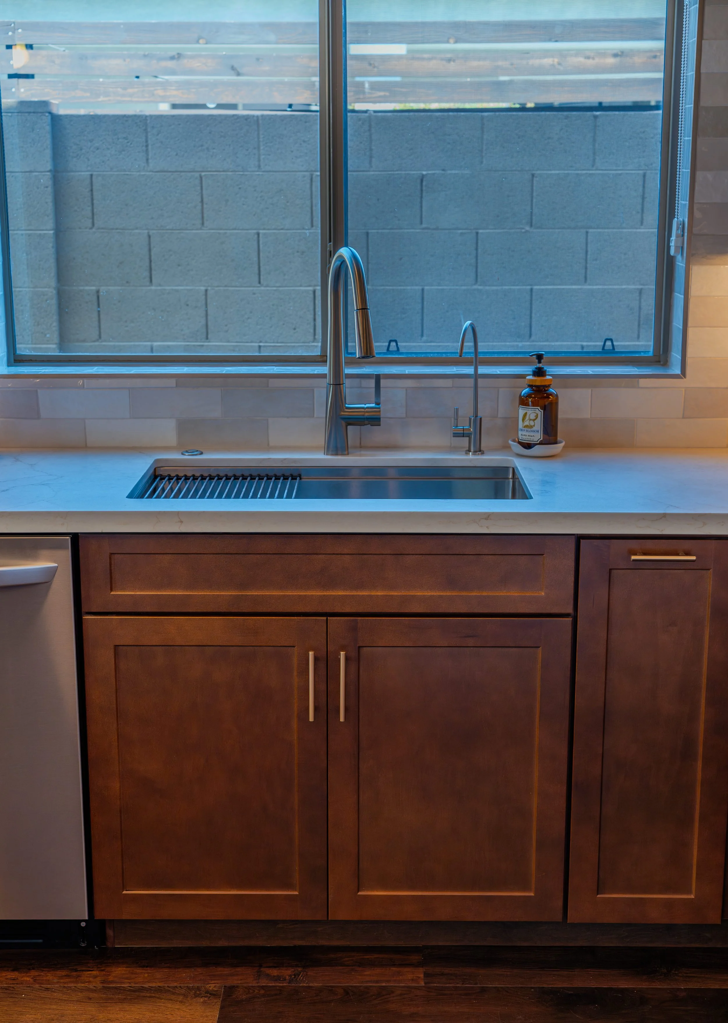 Kitchen sink with brown wooden cabinets, a silver faucet, a soap dispenser, and a large window above the sink with a view of a brick wall and wooden fence outside.