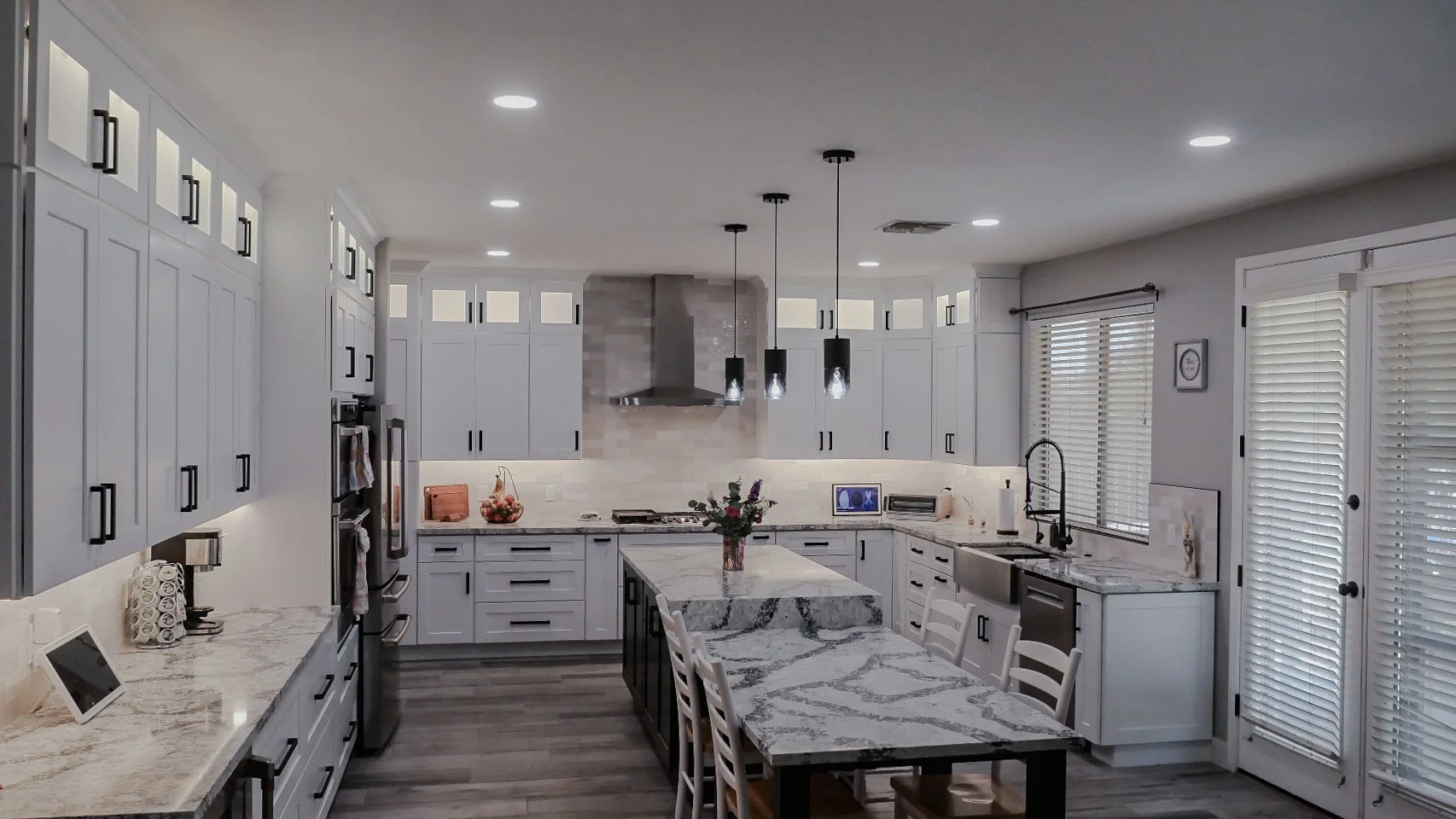 Modern kitchen with white cabinets, marble countertops, a kitchen island, stainless steel appliances, and wooden chairs around the island.