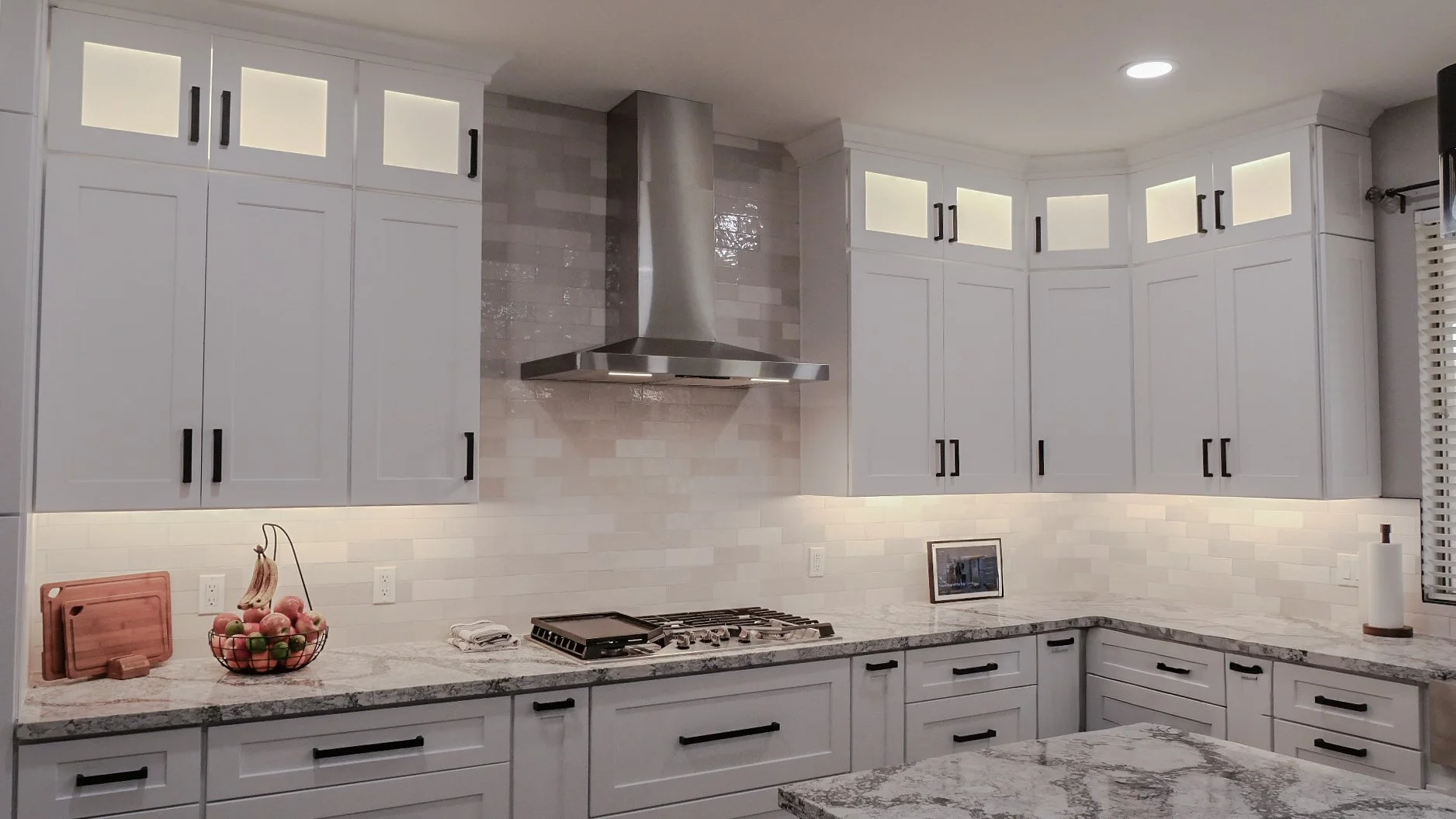 White kitchen cabinets with black handles, granite countertops, a stainless steel range hood, a small framed picture, a fruit basket, a paper towel roll, and a small kitchen appliance.