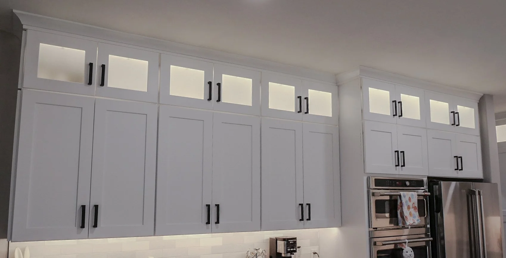 White kitchen cabinets with black handles and upper glass-front doors, stainless steel oven and refrigerator, and a white tile backsplash.