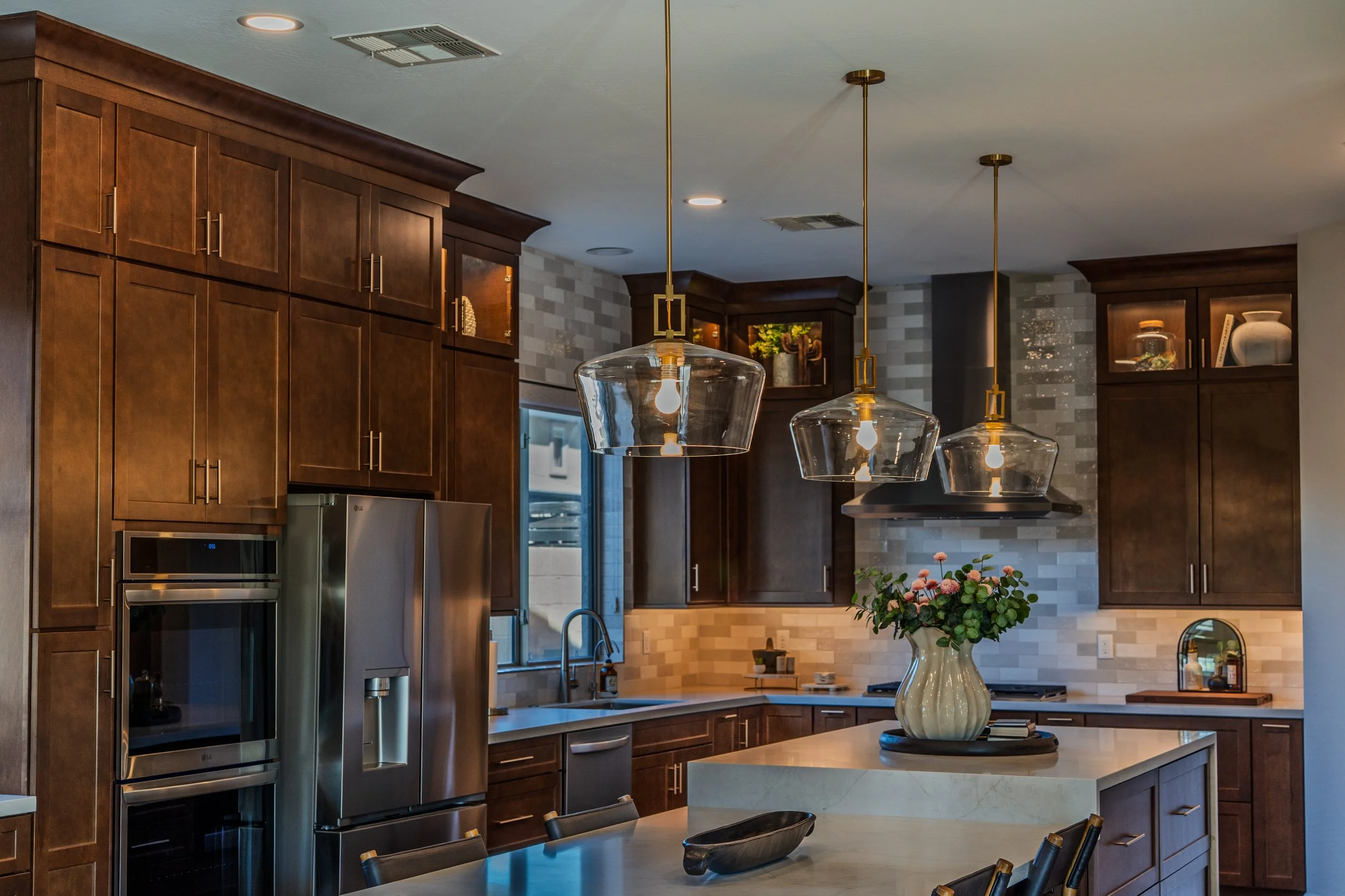 A modern kitchen with brown wooden cabinets, a white marble island with a large cream-colored vase with pink flowers, hanging glass pendant lights, stainless steel appliances, a tiled backsplash, and a small rectangular mirror on the wall.