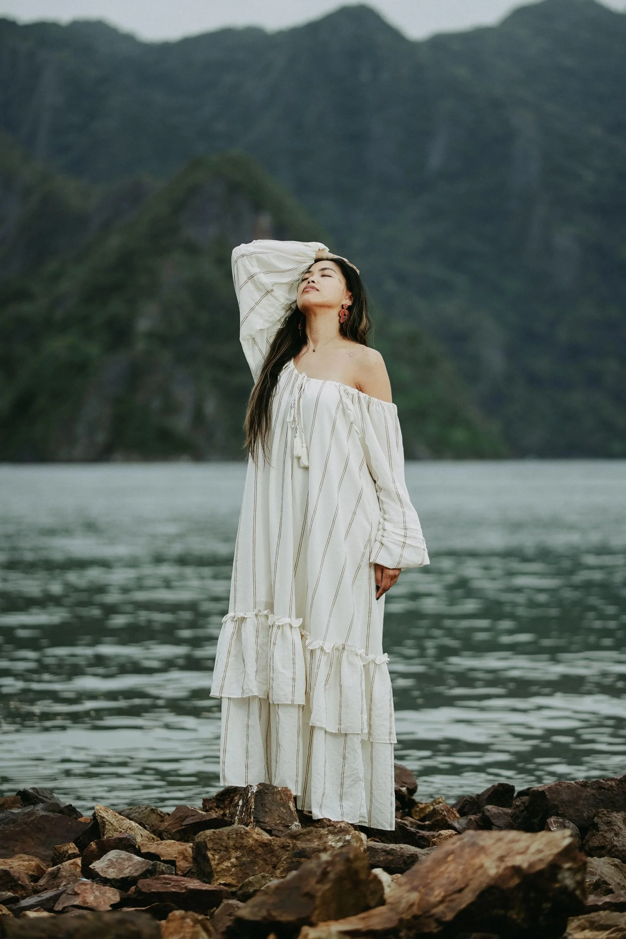 A woman standing on rocks near a body of water with mountains in the background, wearing a white off-the-shoulder dress and red earrings with her eyes closed.