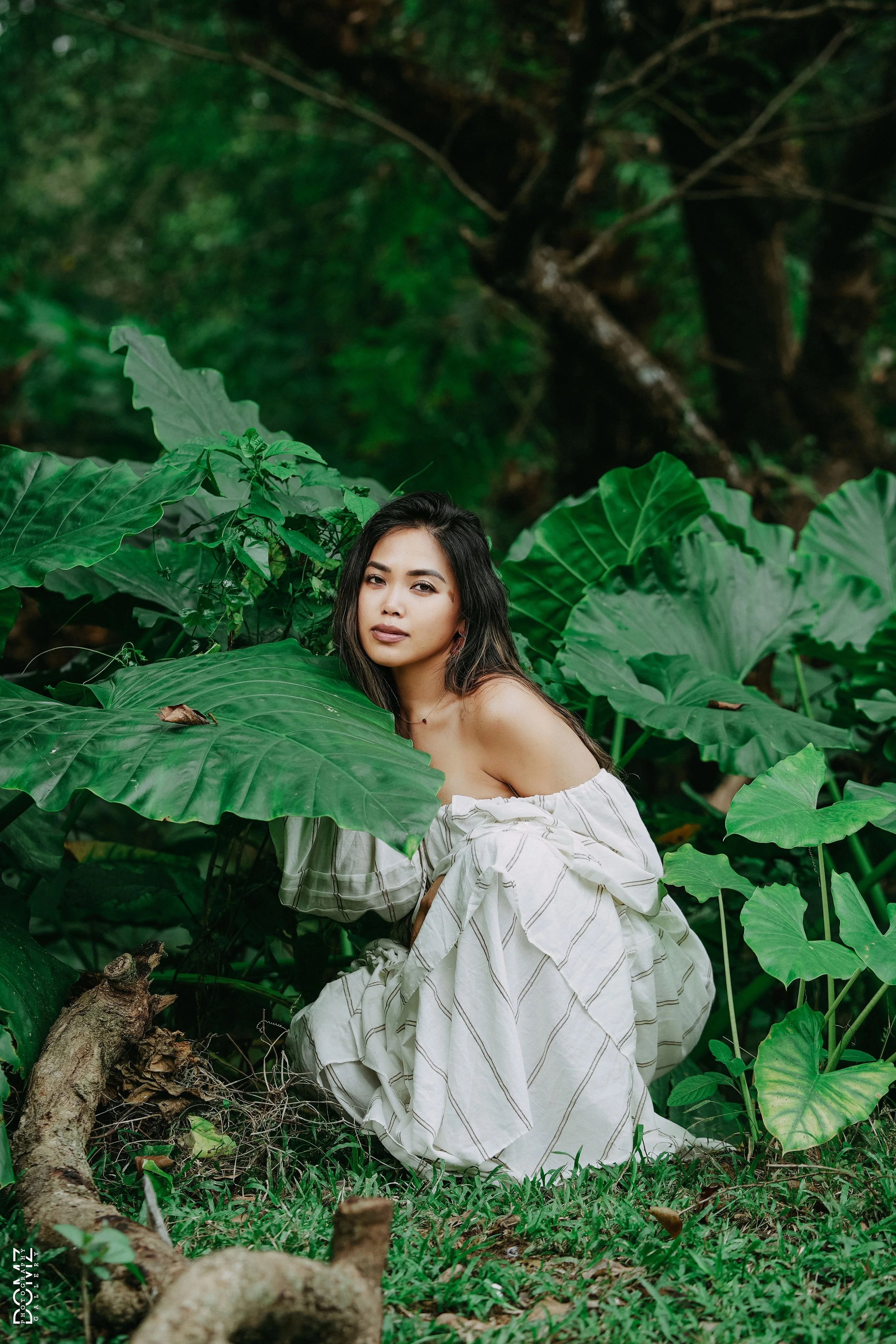 A woman with long dark hair and a neutral expression, wearing an off-shoulder white dress with thin stripes, posing amidst lush green tropical plants and foliage in a forest setting.