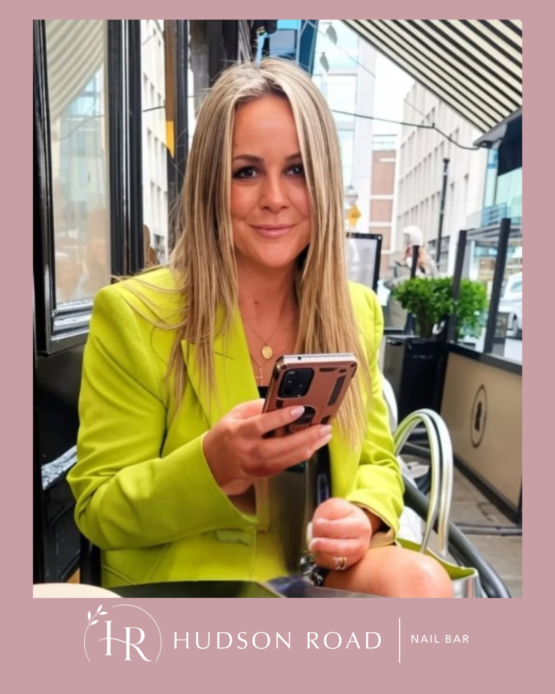 A woman with blonde hair wearing a bright yellow blazer, sitting inside a nail bar, looking at her smartphone, with city buildings and outdoor seating visible through the windows.