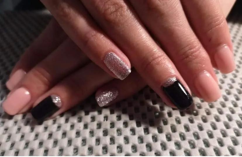 Close-up of hands with manicured nails featuring black, glittery, and nude-colored polish, resting on a textured surface.