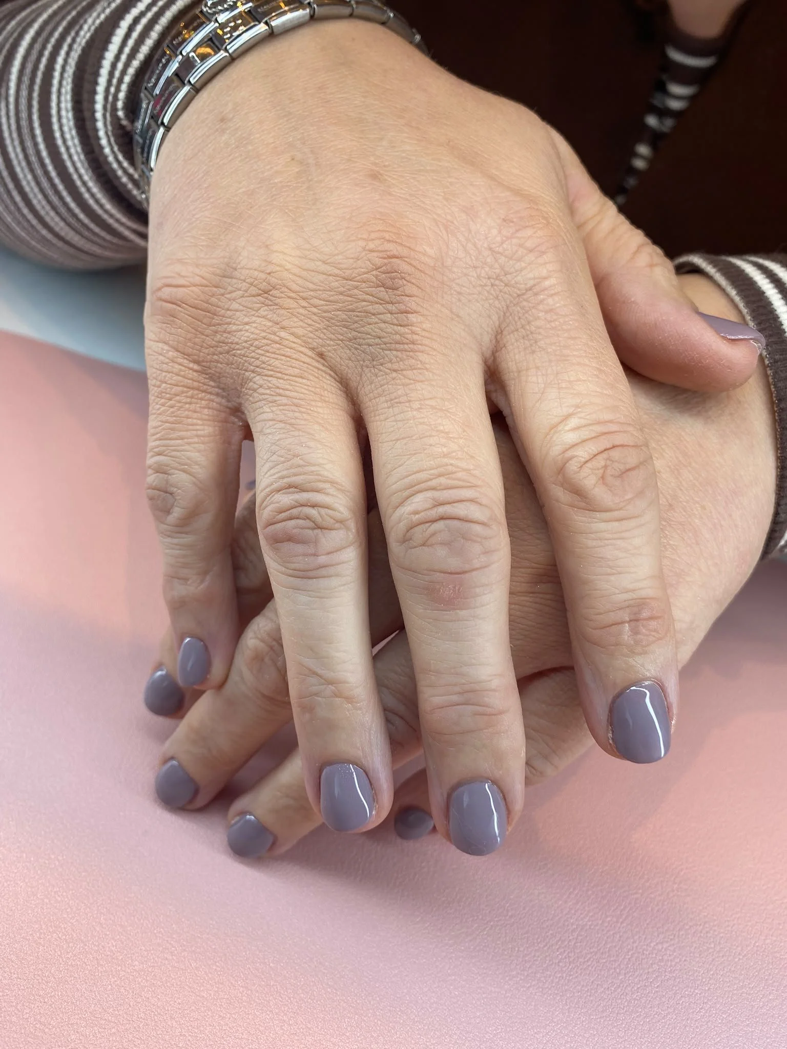 Two hands resting on a pink surface, with nails painted gray and a silver bracelet on one wrist. The person is wearing a brown and white striped long sleeve shirt.