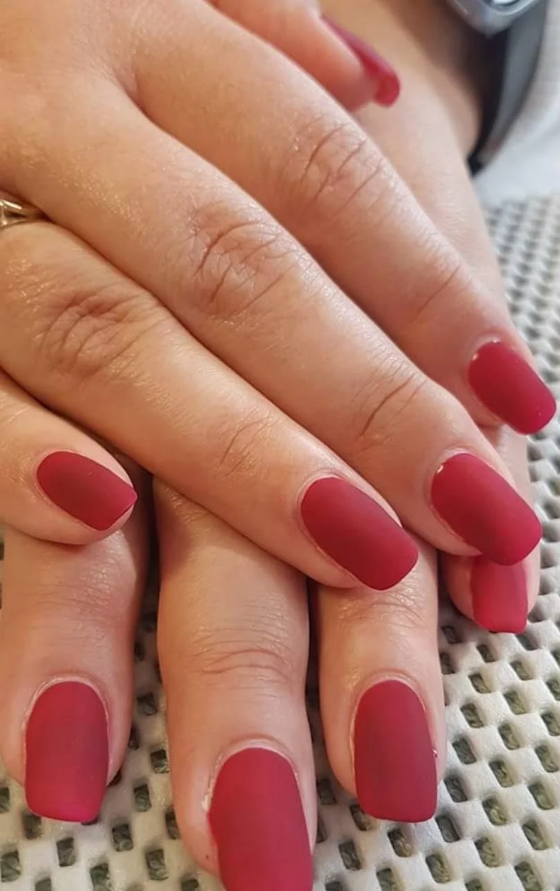 Close-up of hands with red matte almond-shaped nails resting on a textured surface.