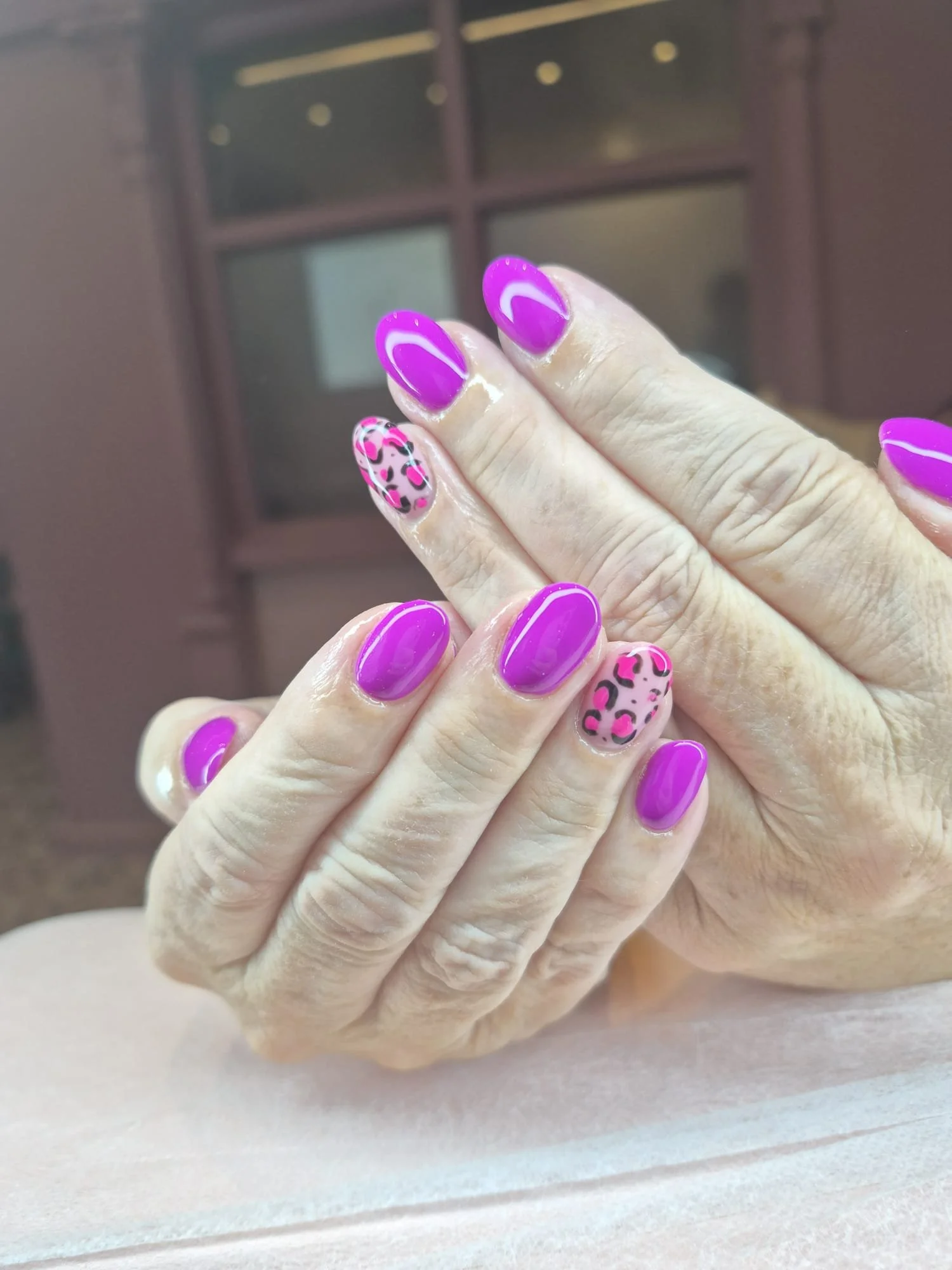 Close-up of a hand with manicured nails painted in purple with white streaks, and one accent nail with a leopard print pattern in pink, black, and white.