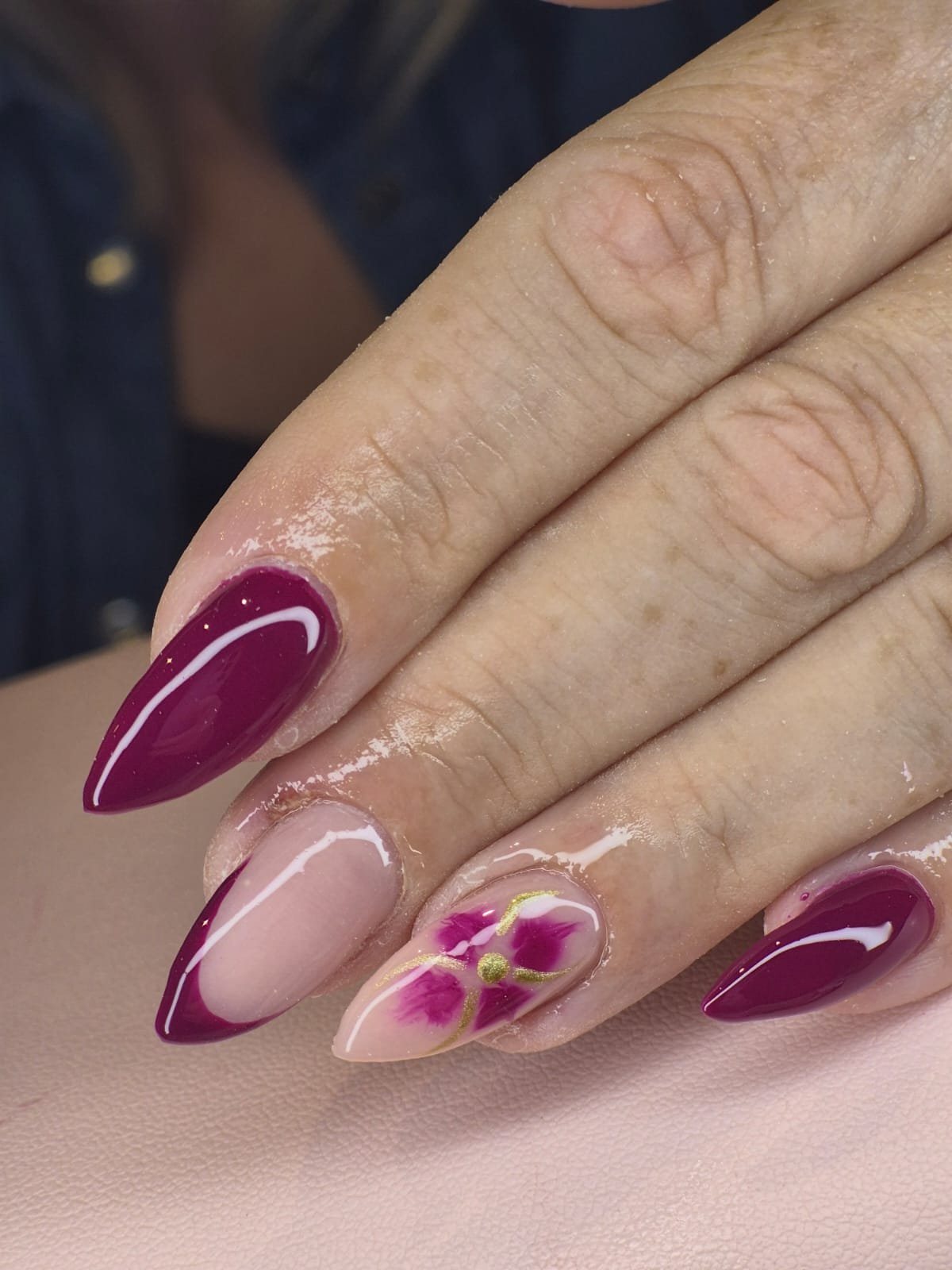 Close-up of a hand showing decorated nails with maroon, pink, and gold nail art, featuring floral and swirl designs.