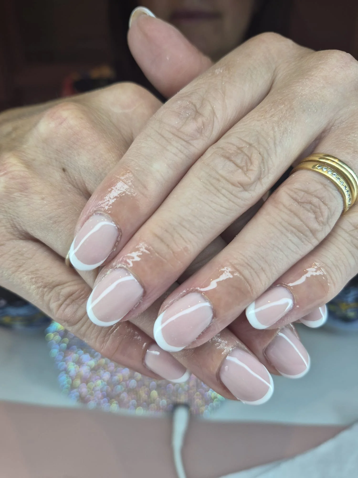 Close-up of a person receiving a manicure with a French nail design, showing multiple rings on their fingers and a background with colorful, shiny dots.