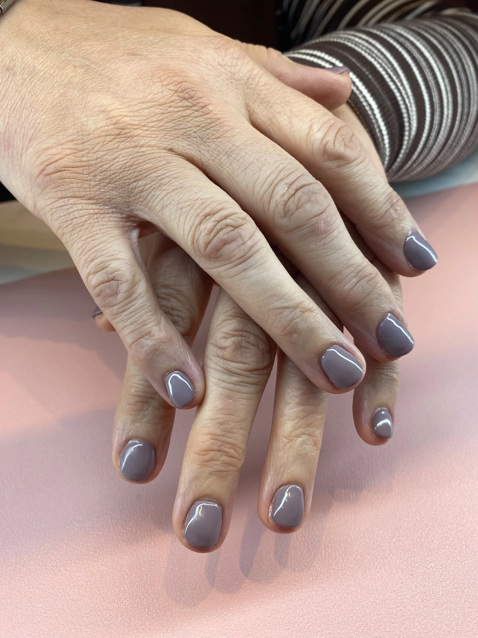 Close-up of a person's hands with gray painted nails, resting on a pink surface. The person is wearing a striped brown and white long-sleeve shirt.