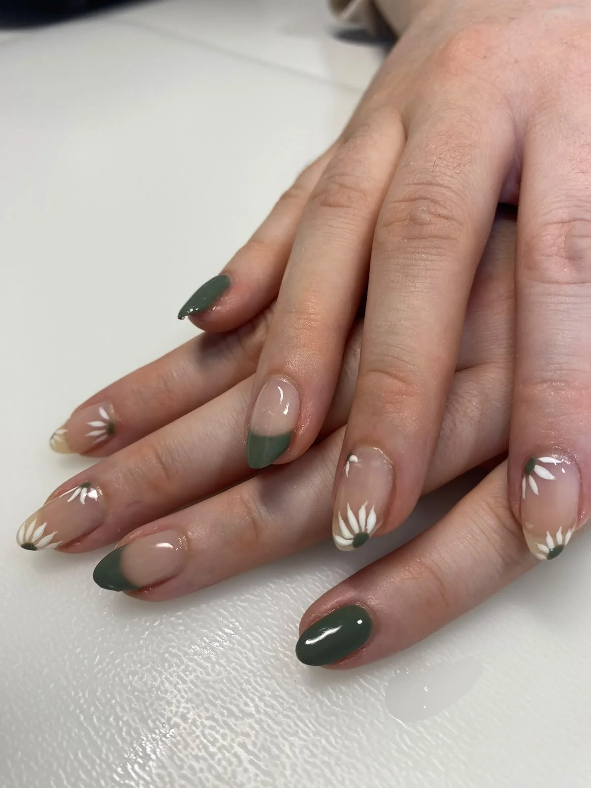 Close-up of a person's hand with manicured nails featuring green tips and white daisy flower designs on a clear base coat.