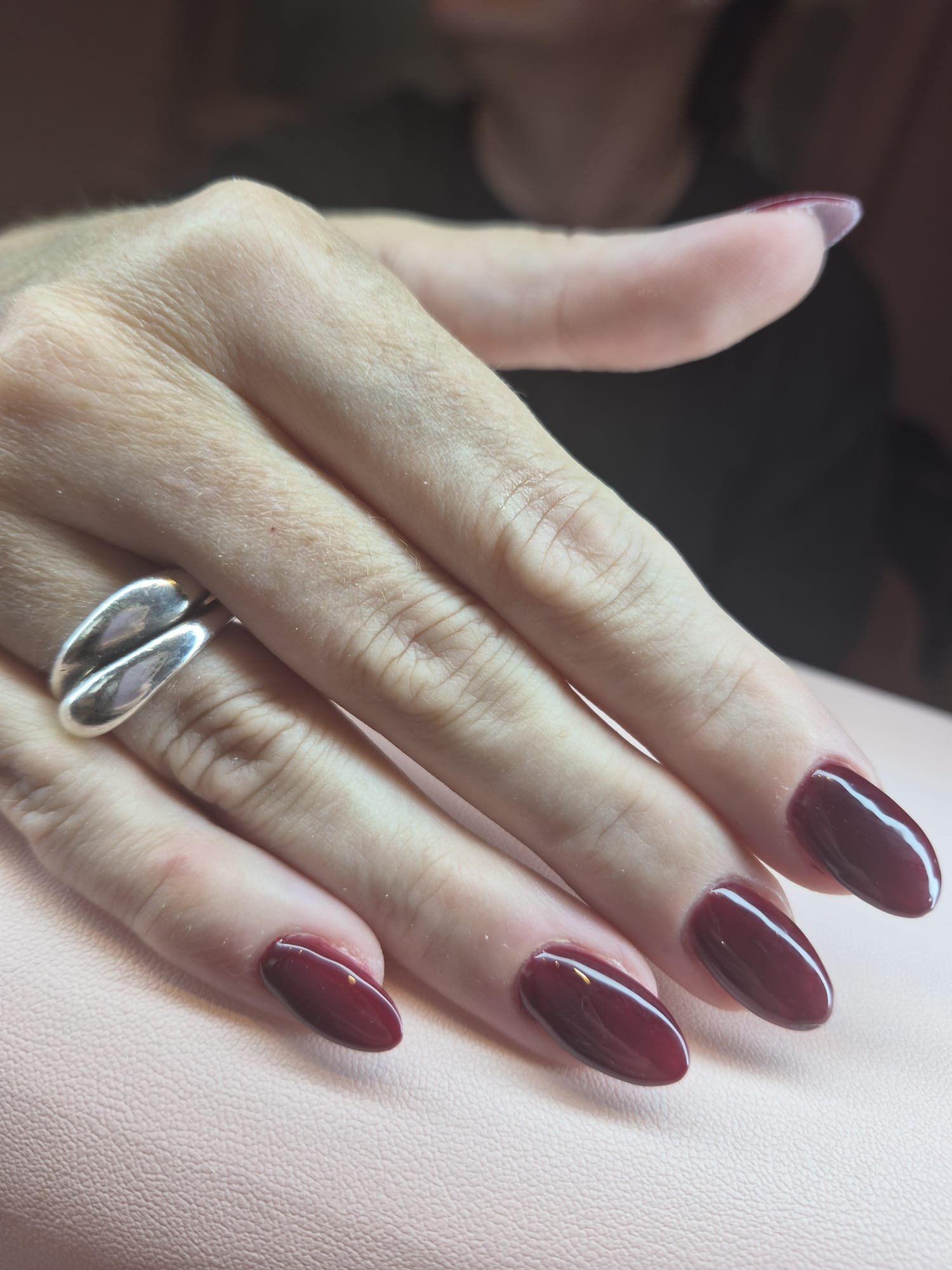 Close-up of a hand with manicured nails painted dark red, wearing a silver ring, resting on a textured beige surface.