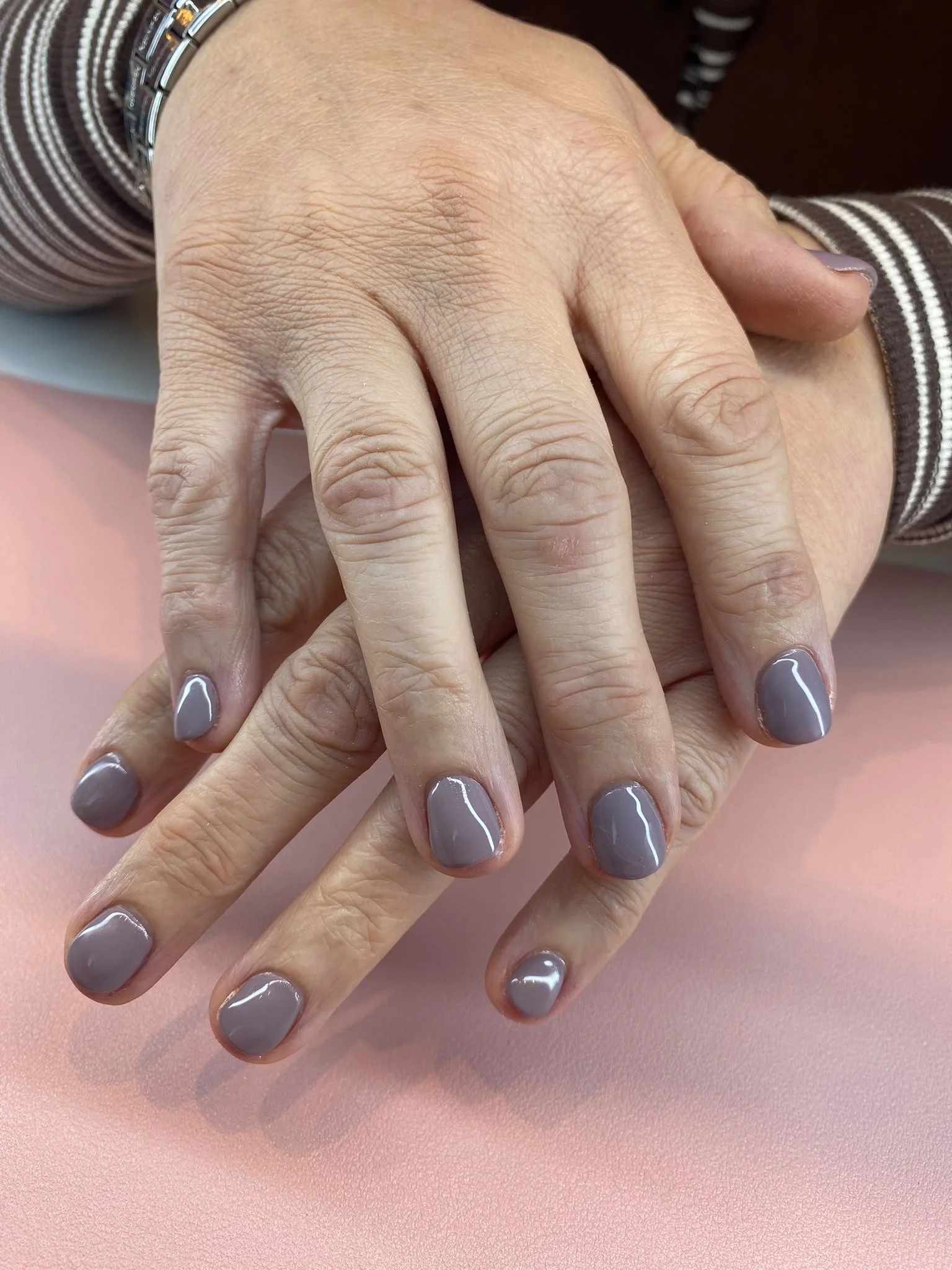 Close-up of a person's hand with manicured nails painted in gray, resting on another person's hand with similar nails, on a pink surface.
