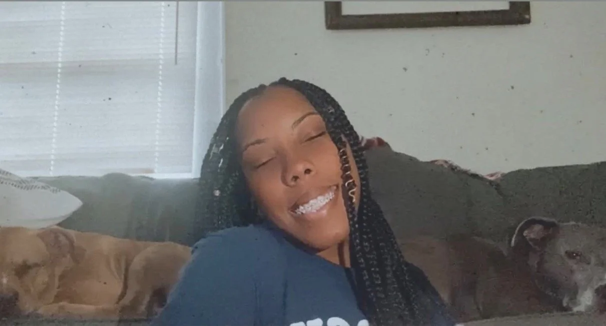 A woman with braided hair smiling with her eyes closed, sitting on a couch with two dogs, one tan and one black, resting beside her.