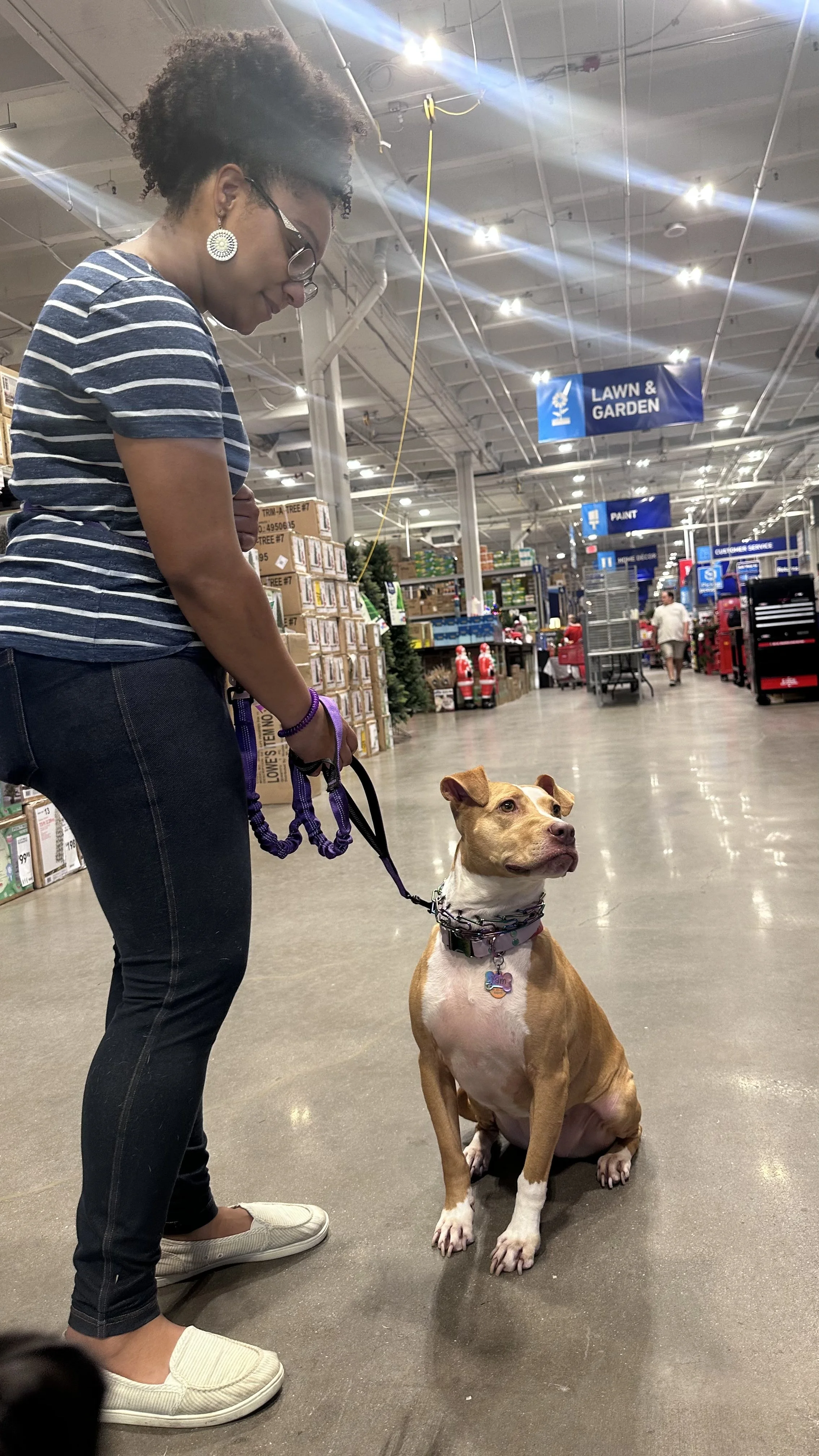 A woman with curly hair, wearing glasses, a striped shirt, and white slip-on shoes, holding a leashed dog sitting on the store's polished floor.