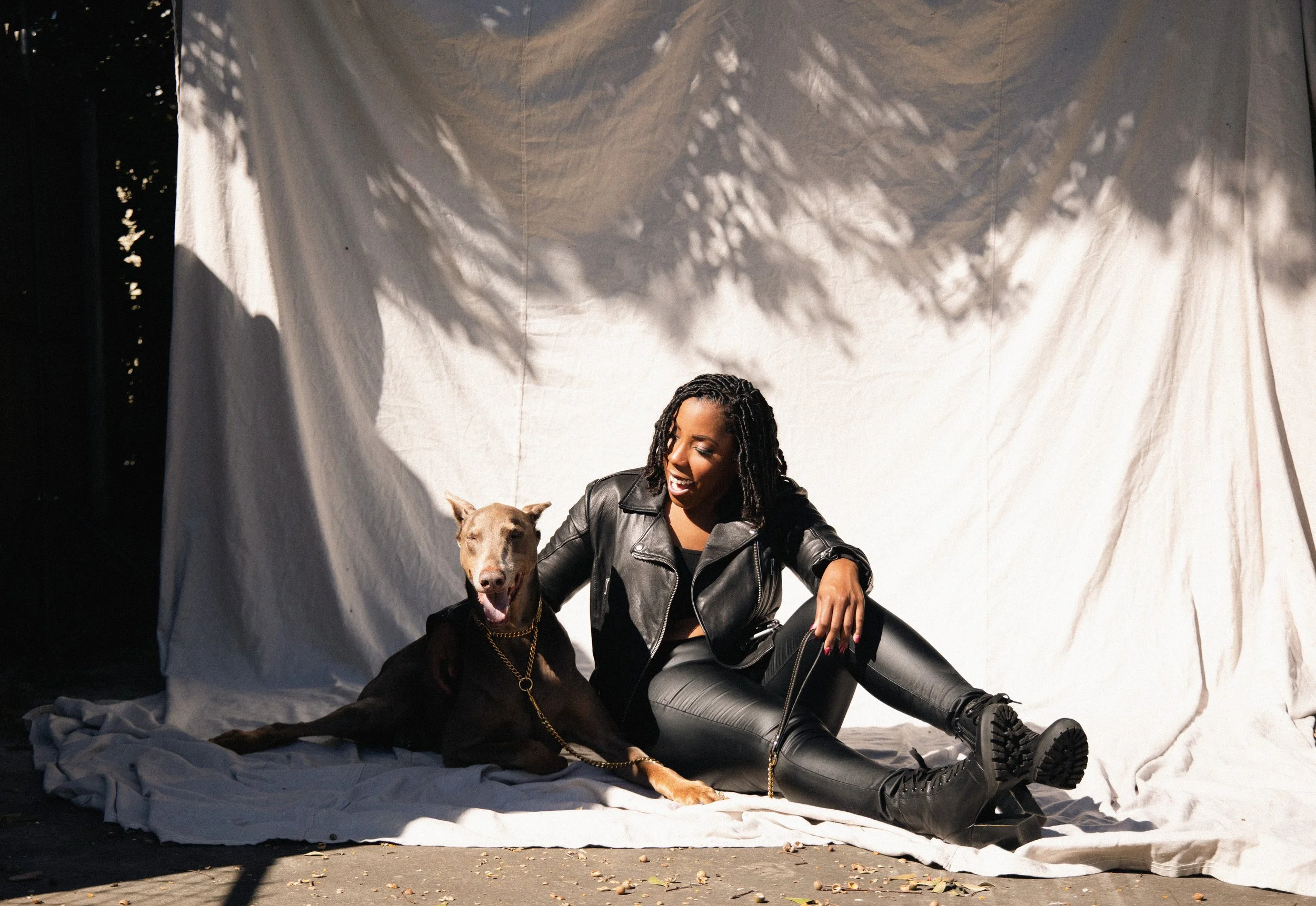 A woman with dreadlocks sitting on a fabric-covered set with a dog, both smiling, in front of a white backdrop with shadows from foliage.
