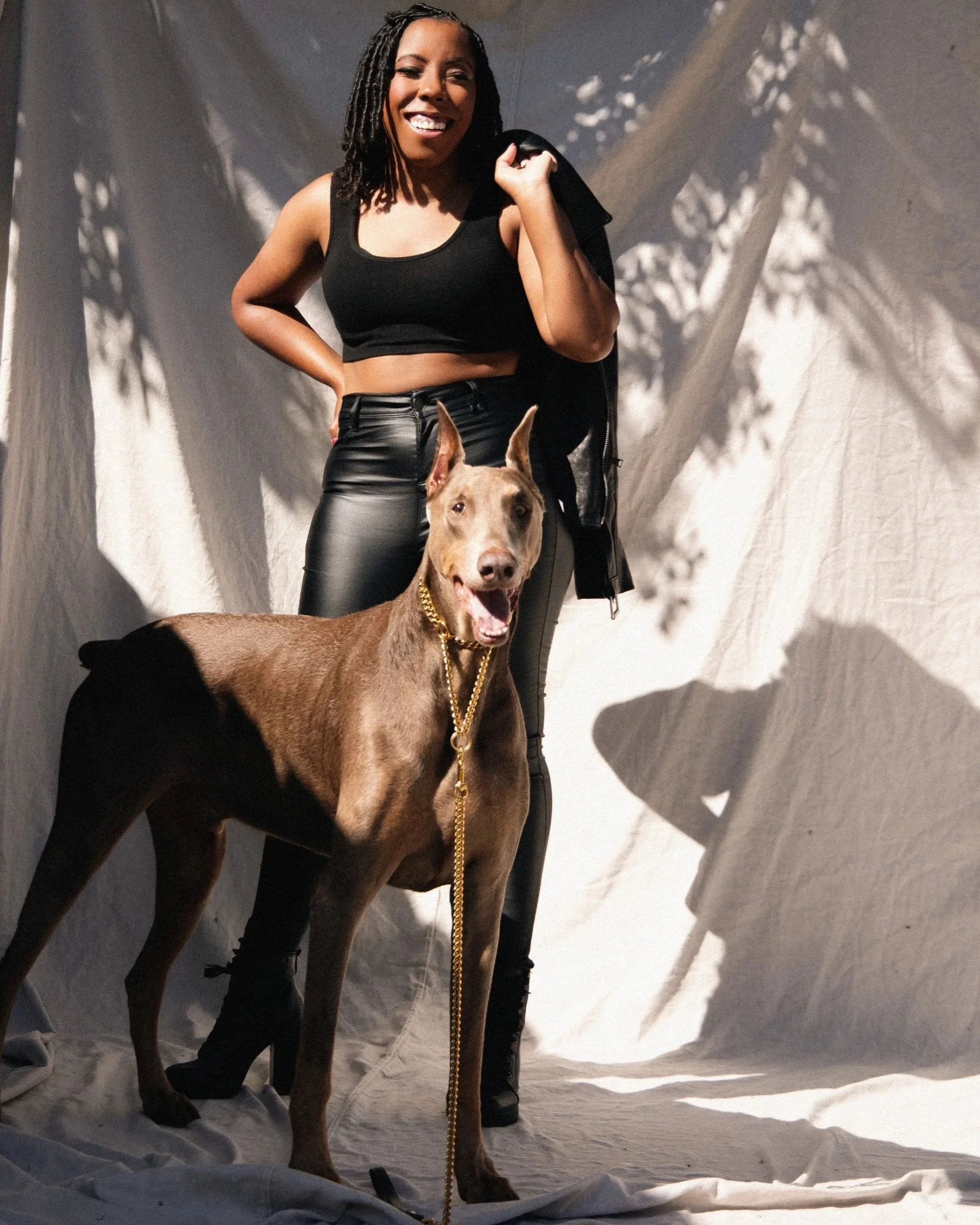 A smiling woman with dreadlocks, wearing a black crop top and leather pants, stands with her hand on her hip holding a black jacket over her shoulder beside a brown dog with a gold chain collar and leash. The setting appears to be a photography studio with white fabric backdrop and shadows cast by lighting.